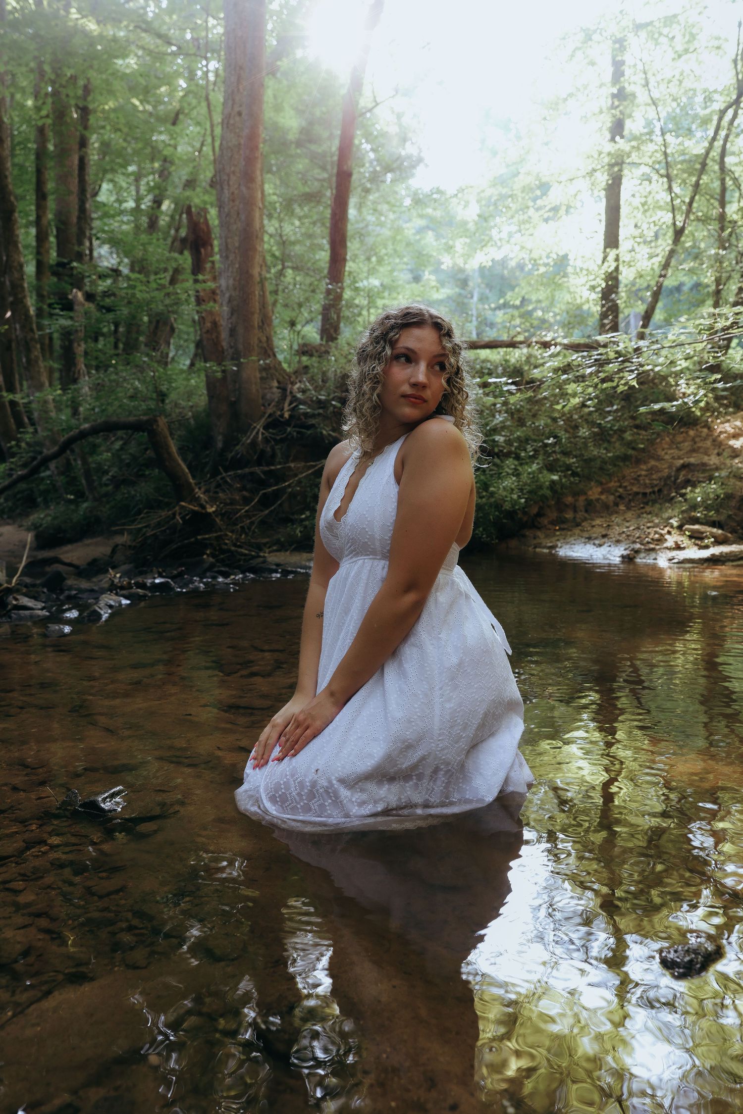 Young woman in a white dress standing in a forest creek with sunlight filtering through trees.