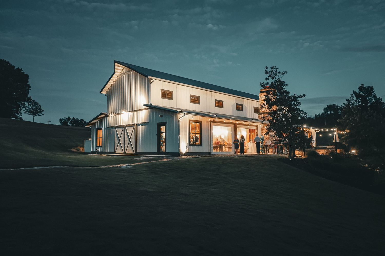 Modern white barn venue glowing at dusk with string lights and sliding doors on a lush green hillside.