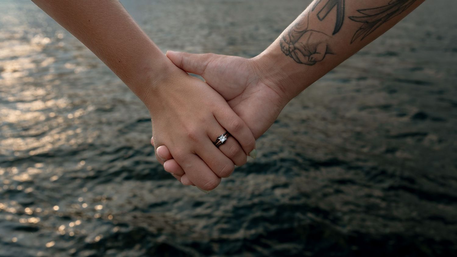 Two people holding hands over water, one with a bird tattoo and a ring, symbolizing love and commitment.