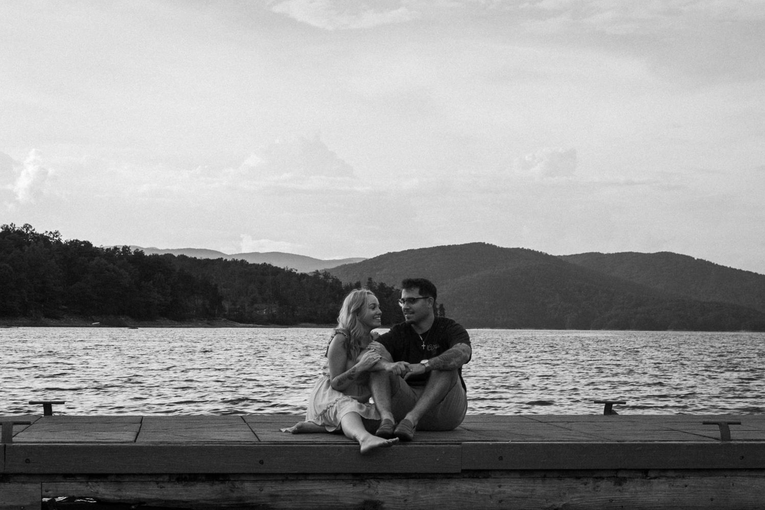 Couple sitting on a dock by a lake with mountain views in black and white.