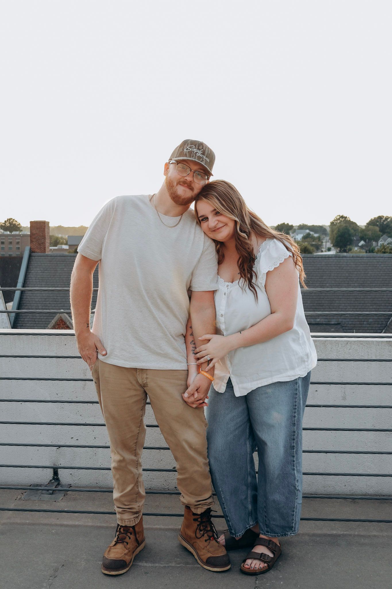 A couple stands together on a rooftop at sunset, woman leaning on mans shoulder affectionately.