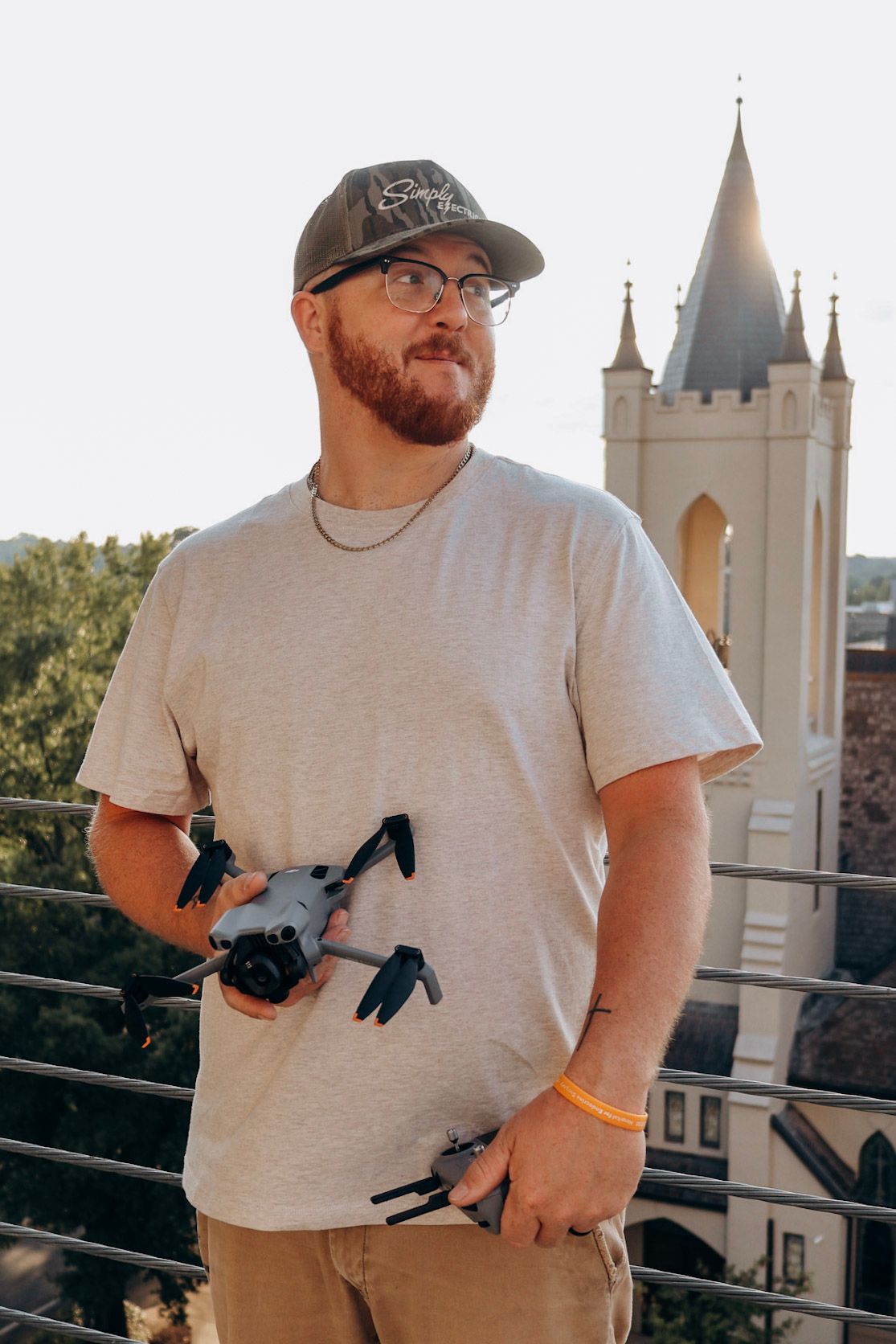 Bearded man in cap and glasses holding a drone on a rooftop with a church steeple behind him.