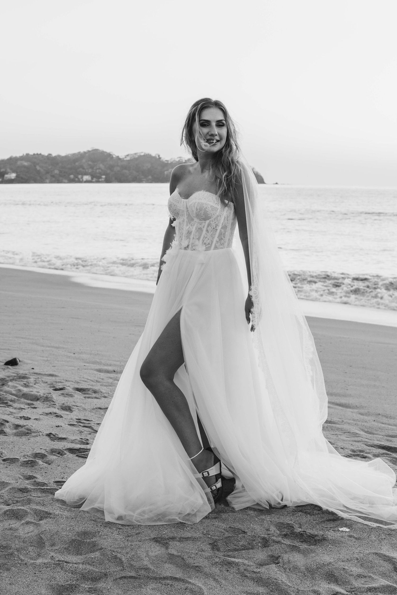 Bride in a strapless lace wedding gown with flowing tulle cape poses on a sandy beach in sayulita