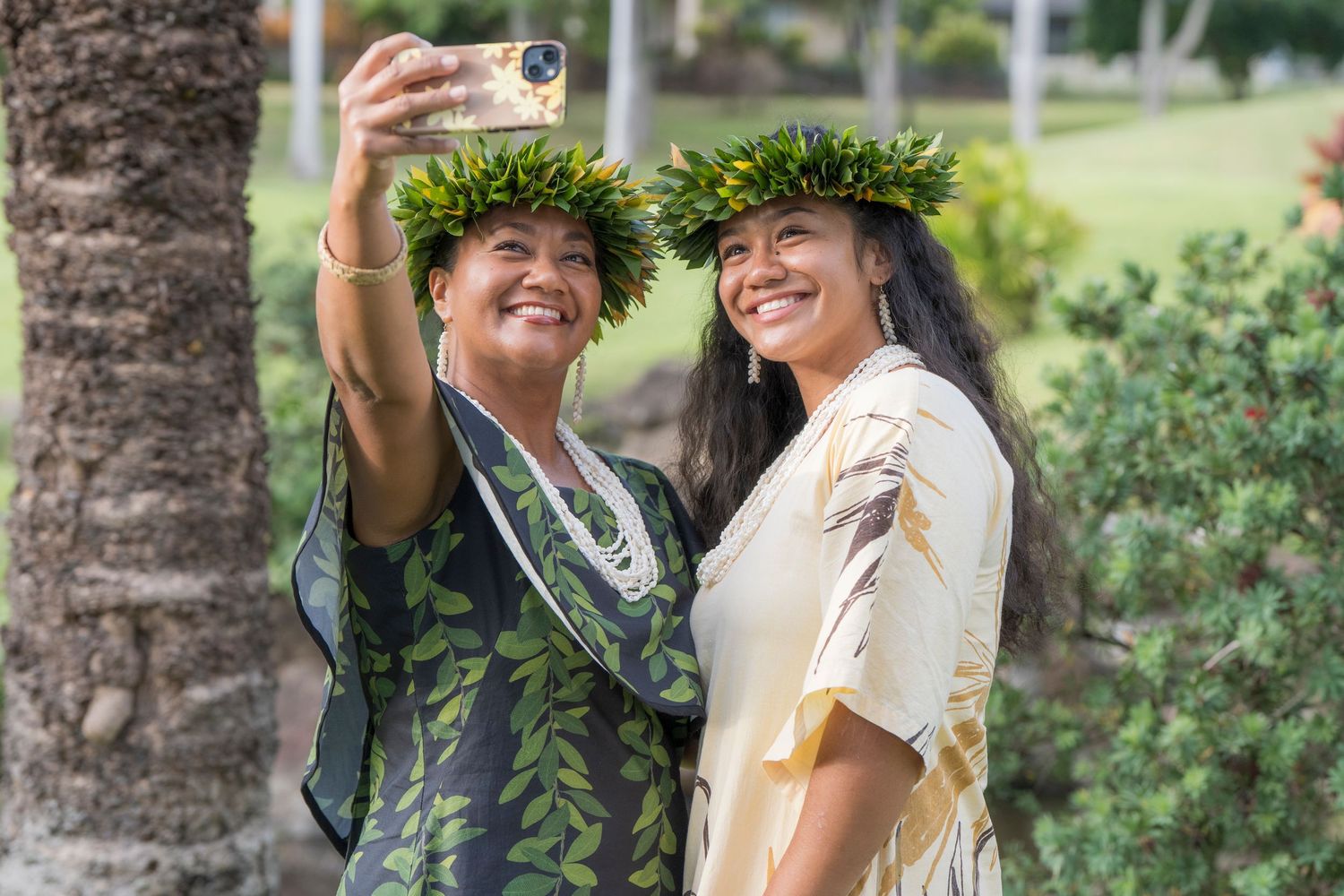 Two women in traditional Hawaiian hula attire with leaf crowns smile while taking a selfie outdoors.
