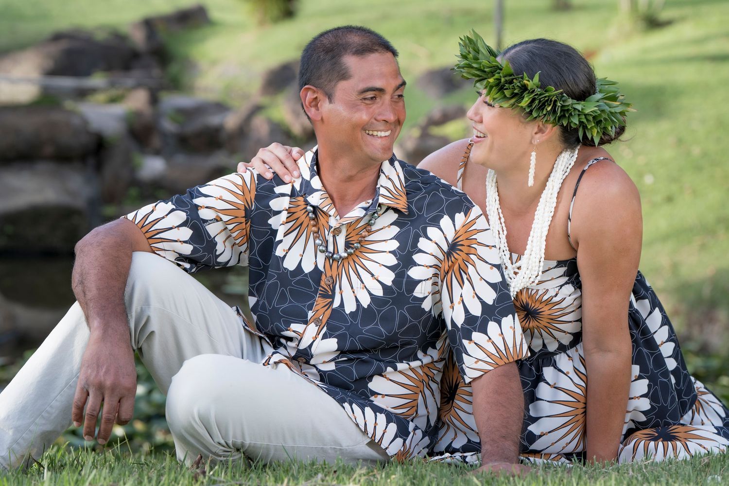 Couple in matching Hawaiian floral outfits sitting on grass, woman wearing a green leaf crown, smiling at each other outdoors.