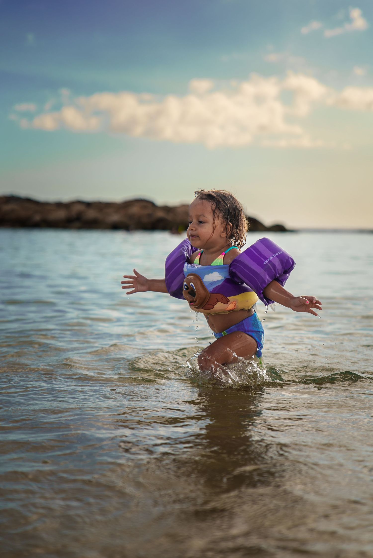 Young child splashing through shallow ocean water in purple floaties