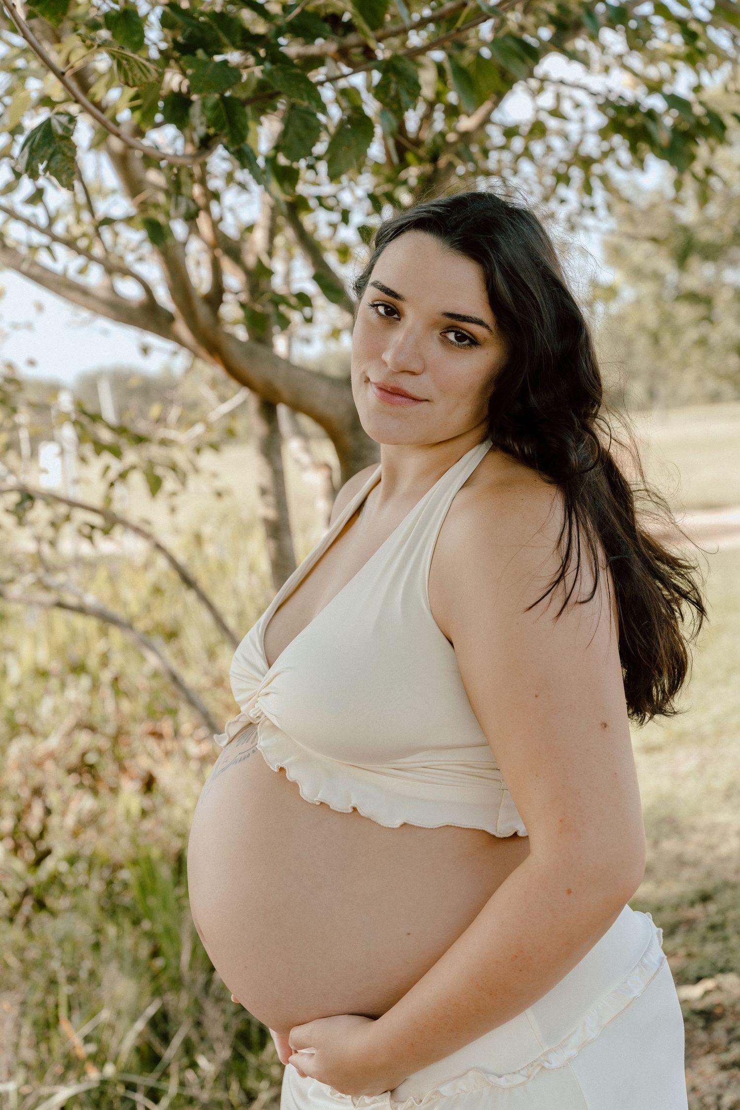 Pregnant woman with dark hair in cream halter top holding baby bump under tree outdoors.
