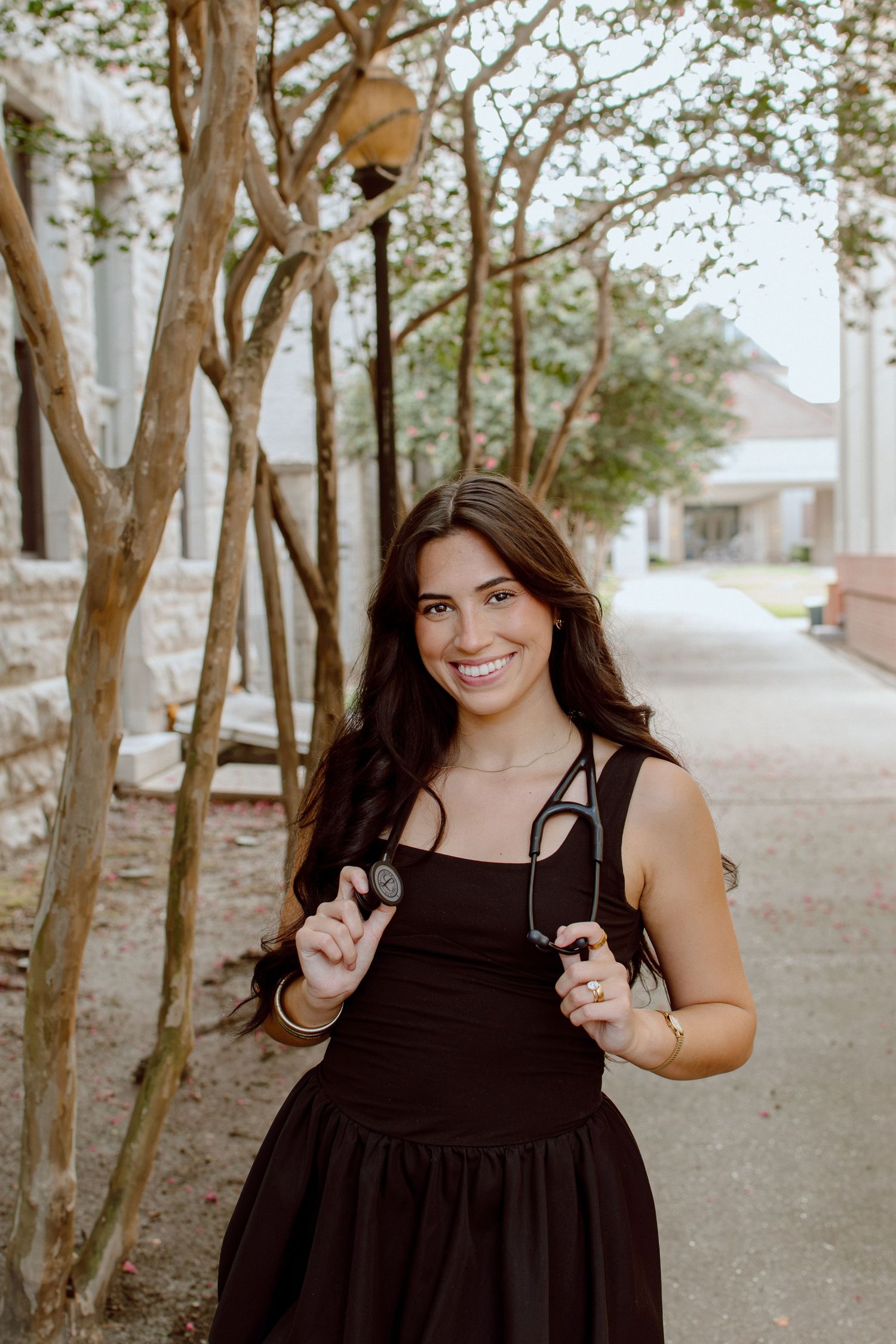 Smiling brunette woman in black dress holding sunglasses outdoors among trees for senior photos.