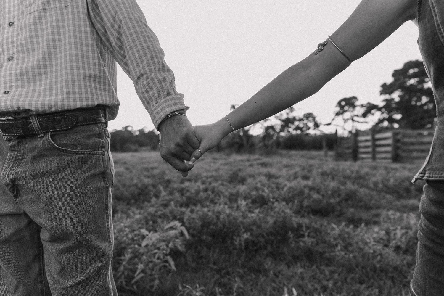 Black and white photo of a couple holding hands in a rural field with a wooden fence in the background.