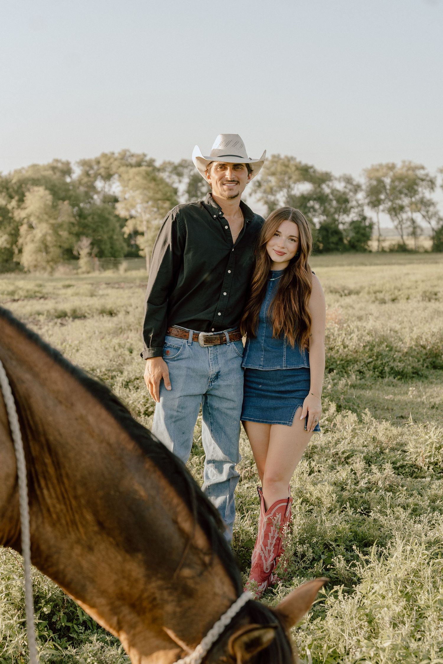 Couple standing in a sunlit green field with a horse in foreground, man in cowboy hat, woman in denim dress.