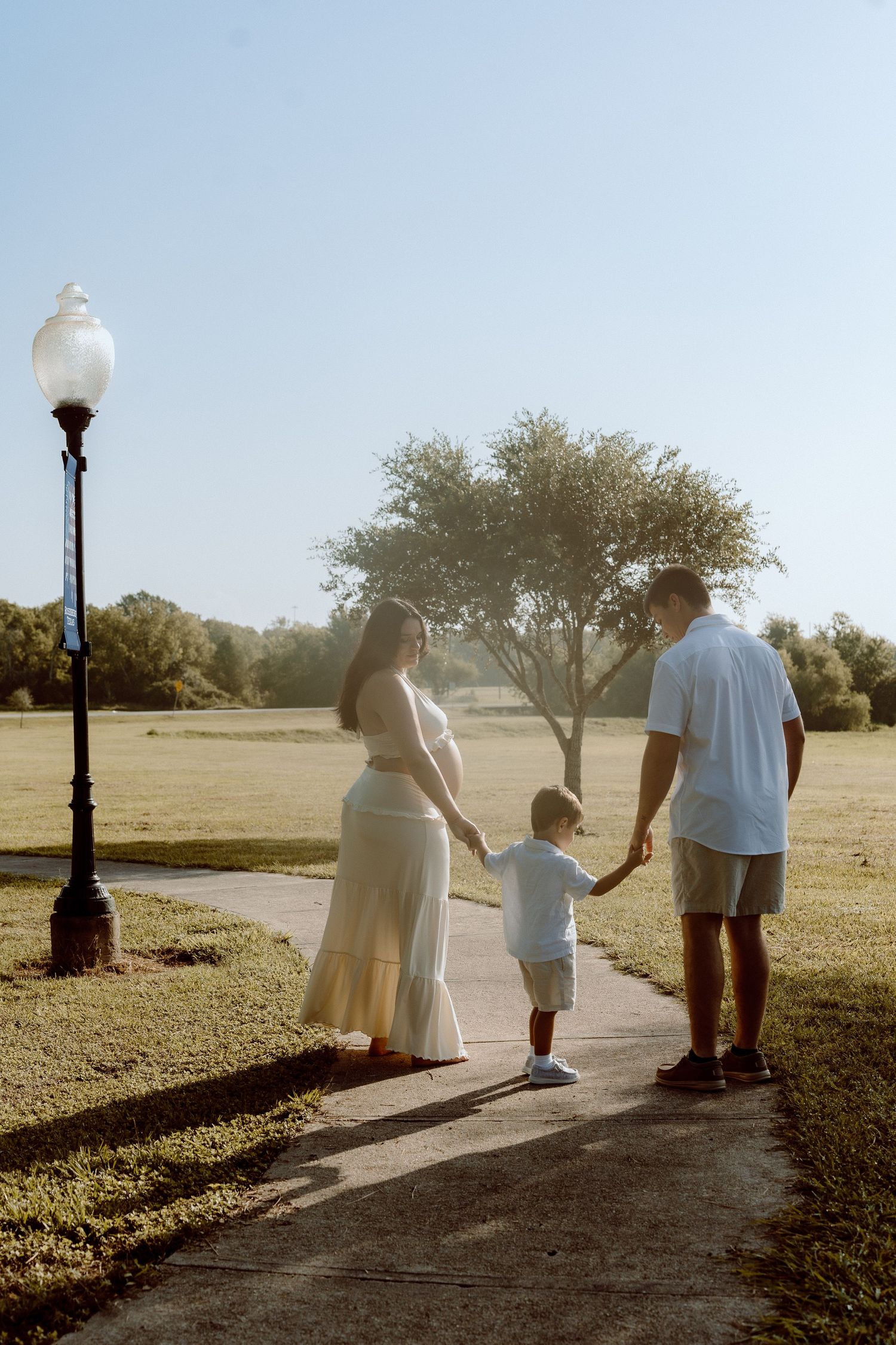 A pregnant woman in white, a man, and a toddler walk hand-in-hand along a sunny park path at golden hour.