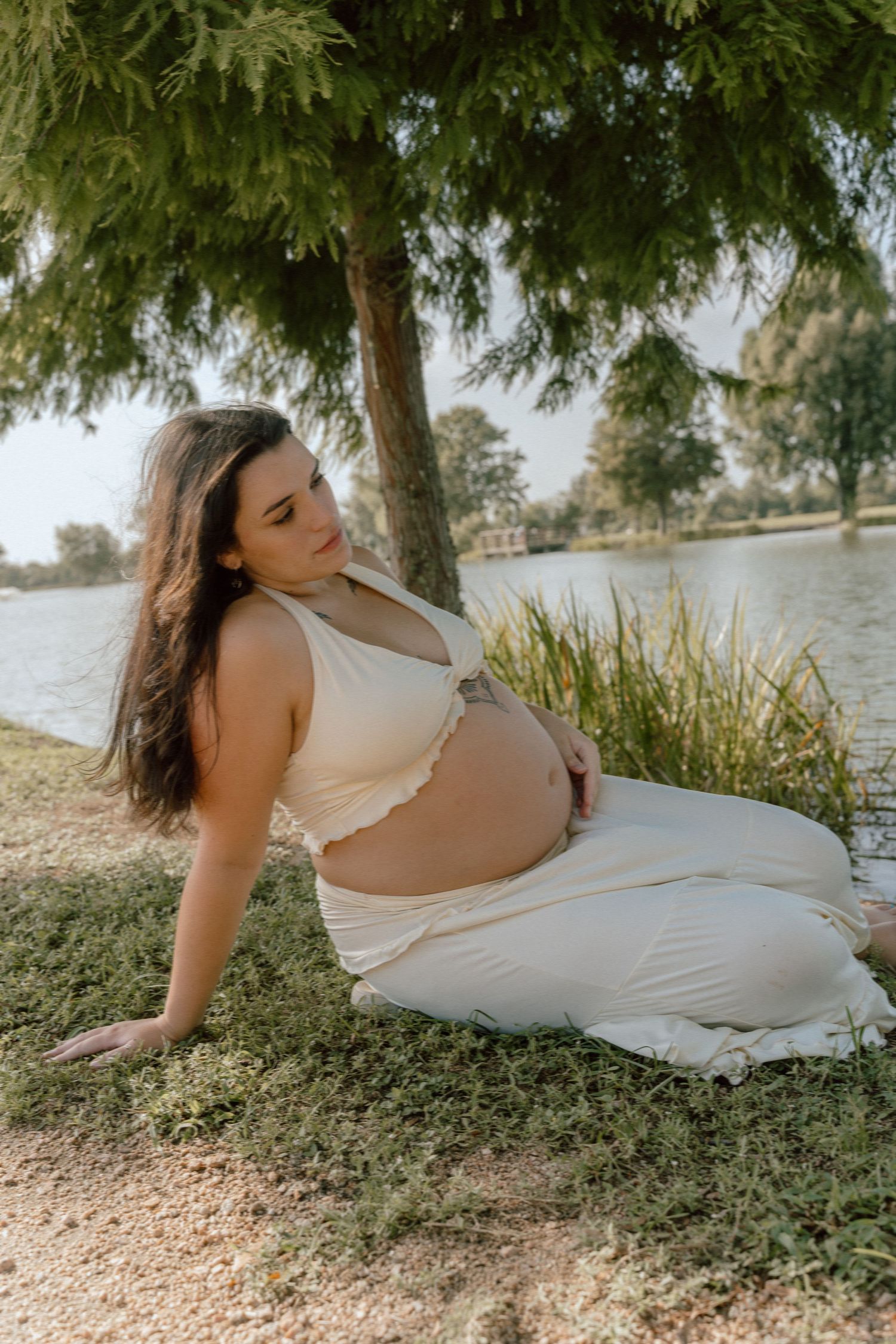 Pregnant woman in white crop top and skirt sitting by lake gazing down at baby bump.