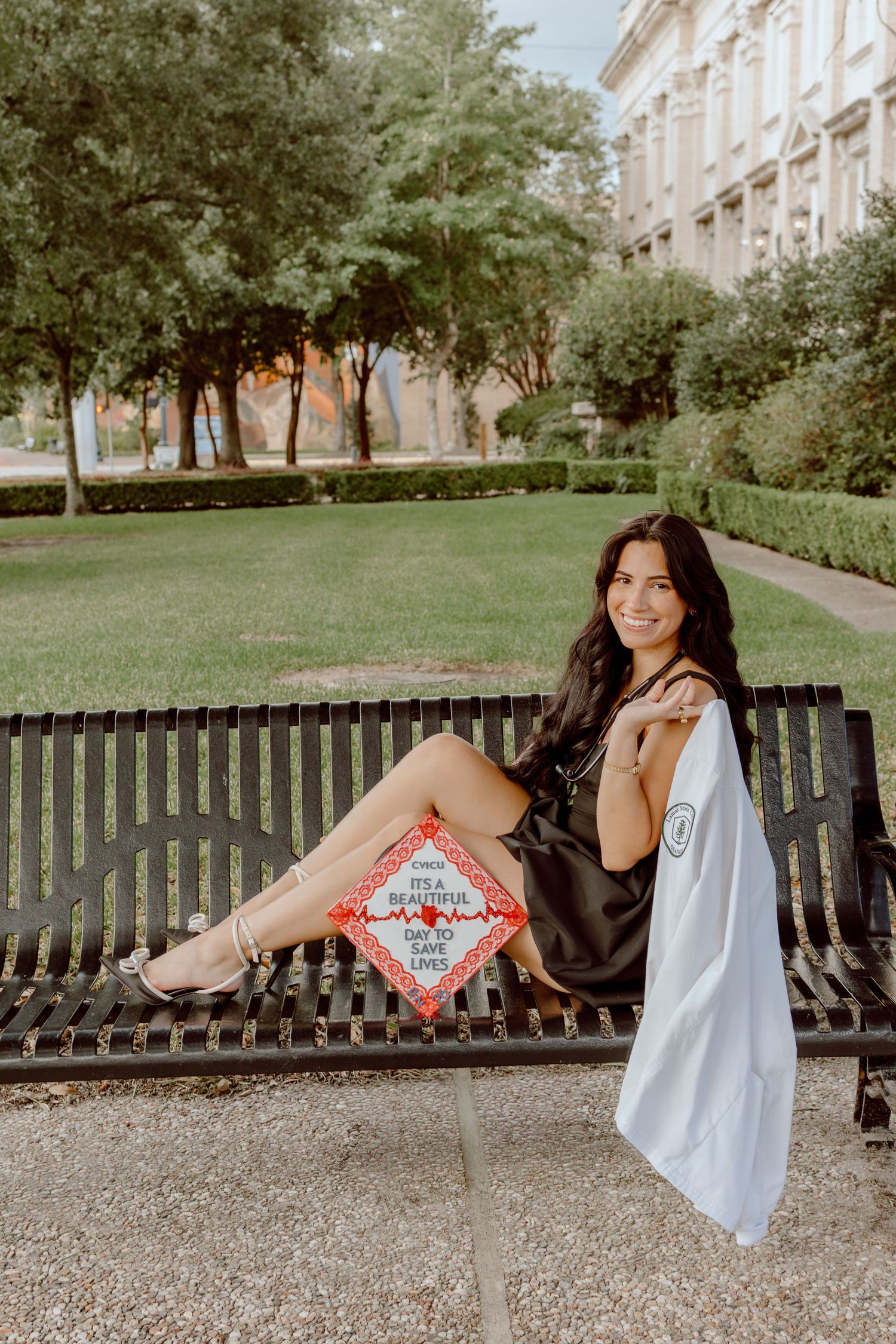 Smiling brunette woman sitting on bench with graduation cap and white gown for senior photos.