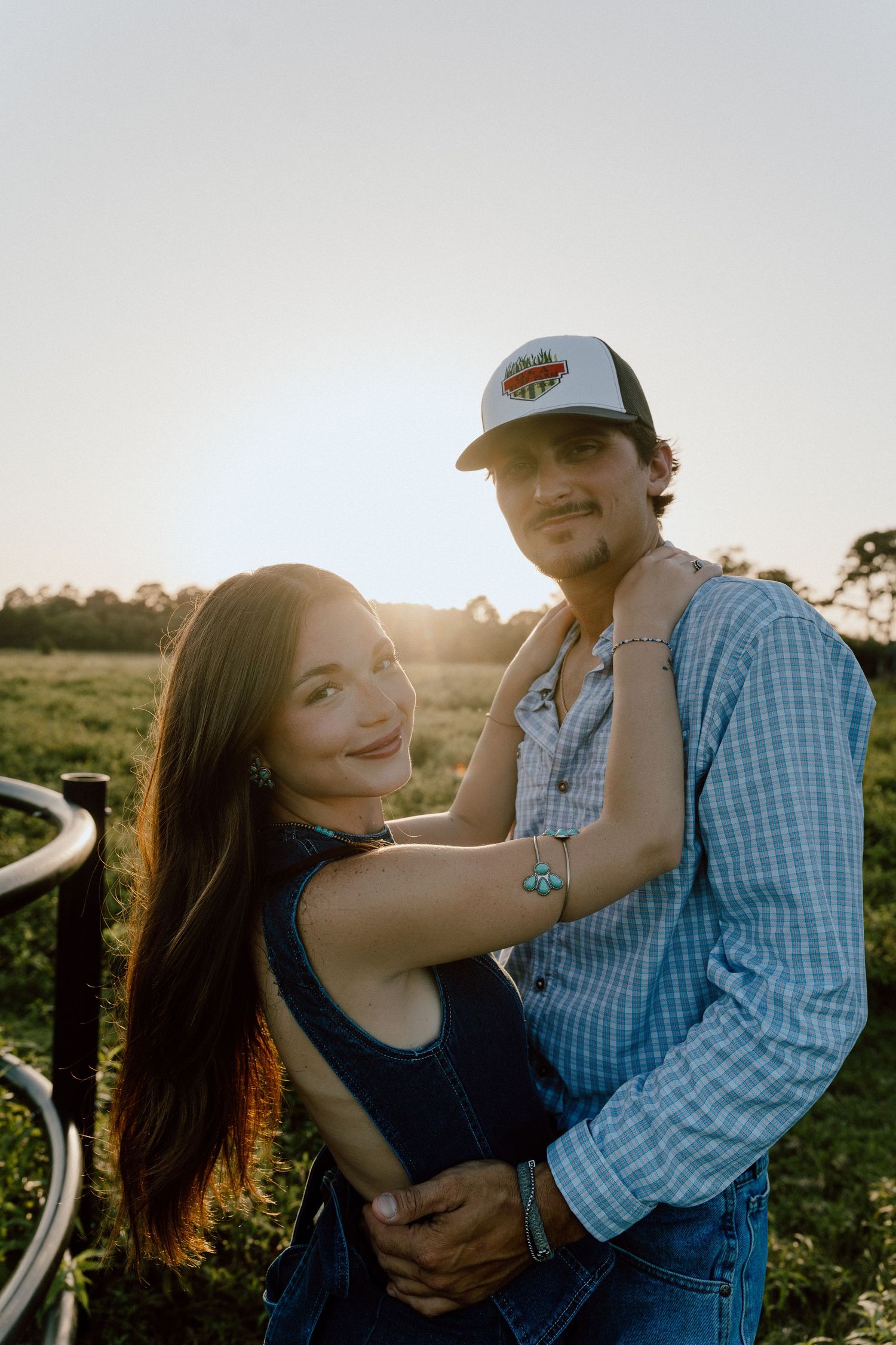 Couple embracing at golden hour near a farm fence, man in trucker hat and plaid shirt, woman in denim.