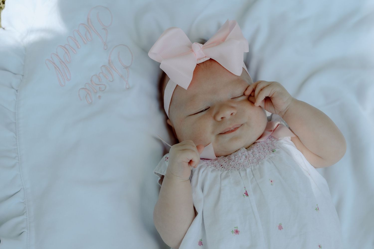 Sleeping newborn baby girl wearing pink bow headband smiling softly on white personalized blanket.