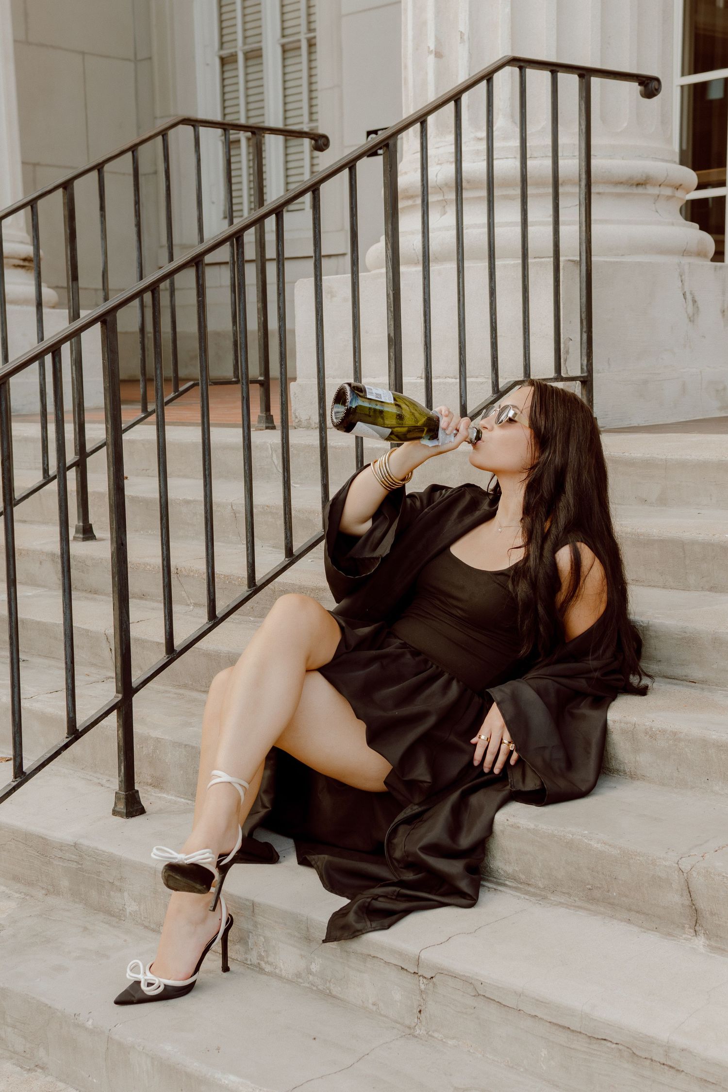 Woman in black dress sitting on marble steps drinking from a wine bottle while wearing silver heels.