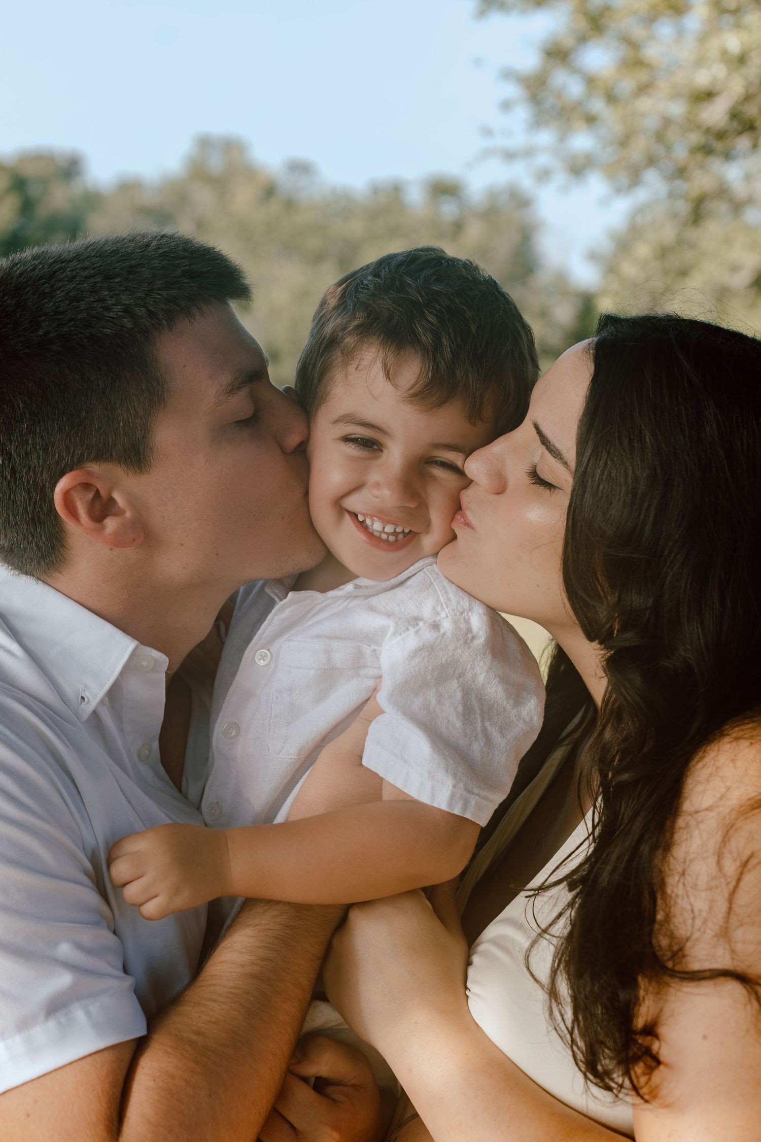 Parents kissing their laughing toddler son on both cheeks in a sunny outdoor setting.