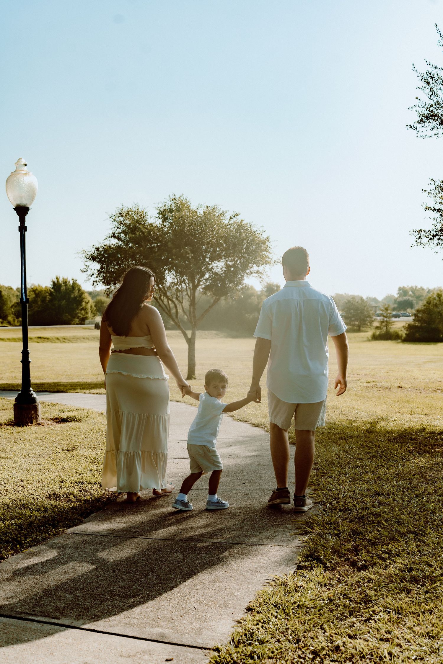 Family of three walking hand in hand on a sunny park path, viewed from behind.