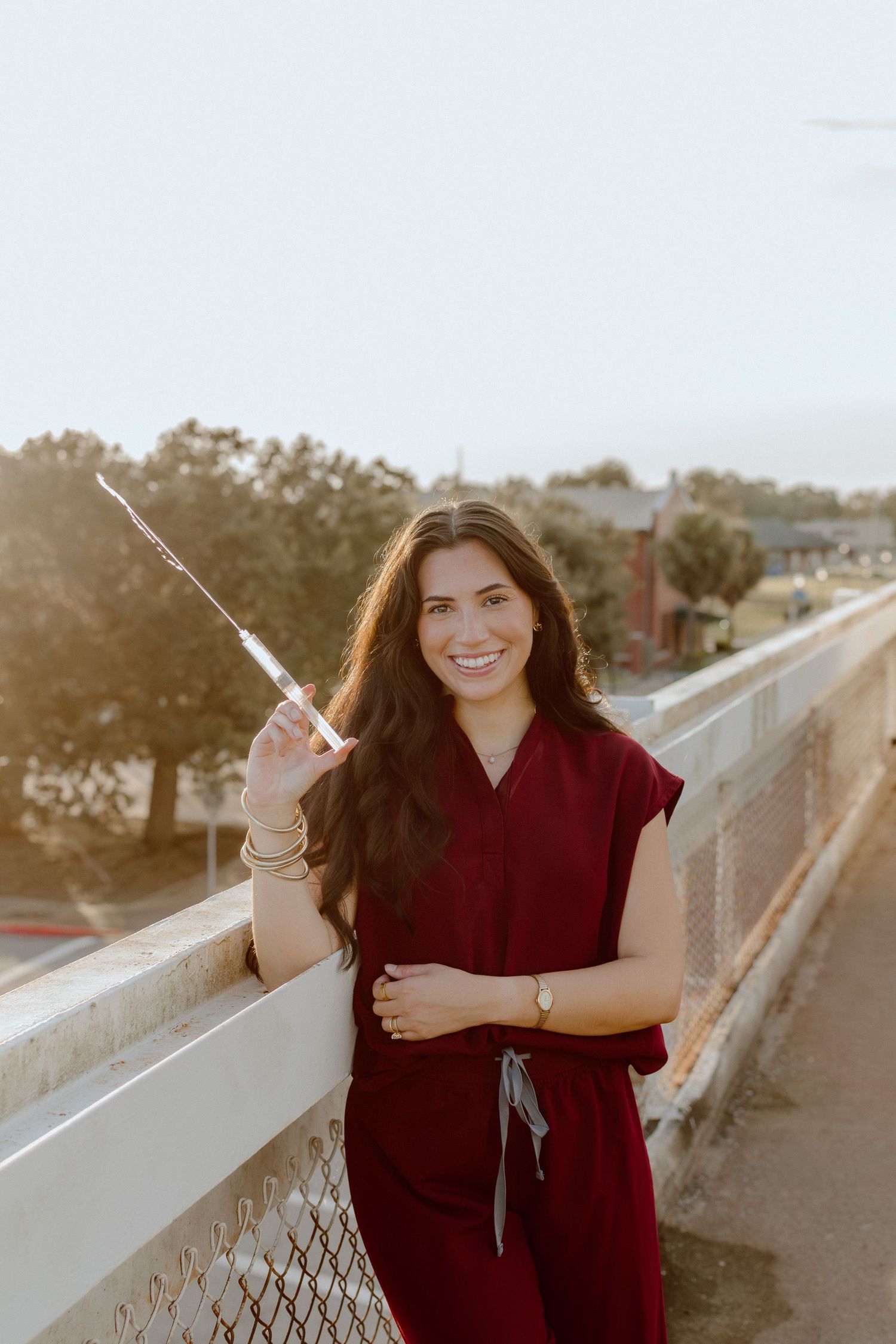 Smiling woman in burgundy scrubs holding a syringe on a bridge at golden hour.