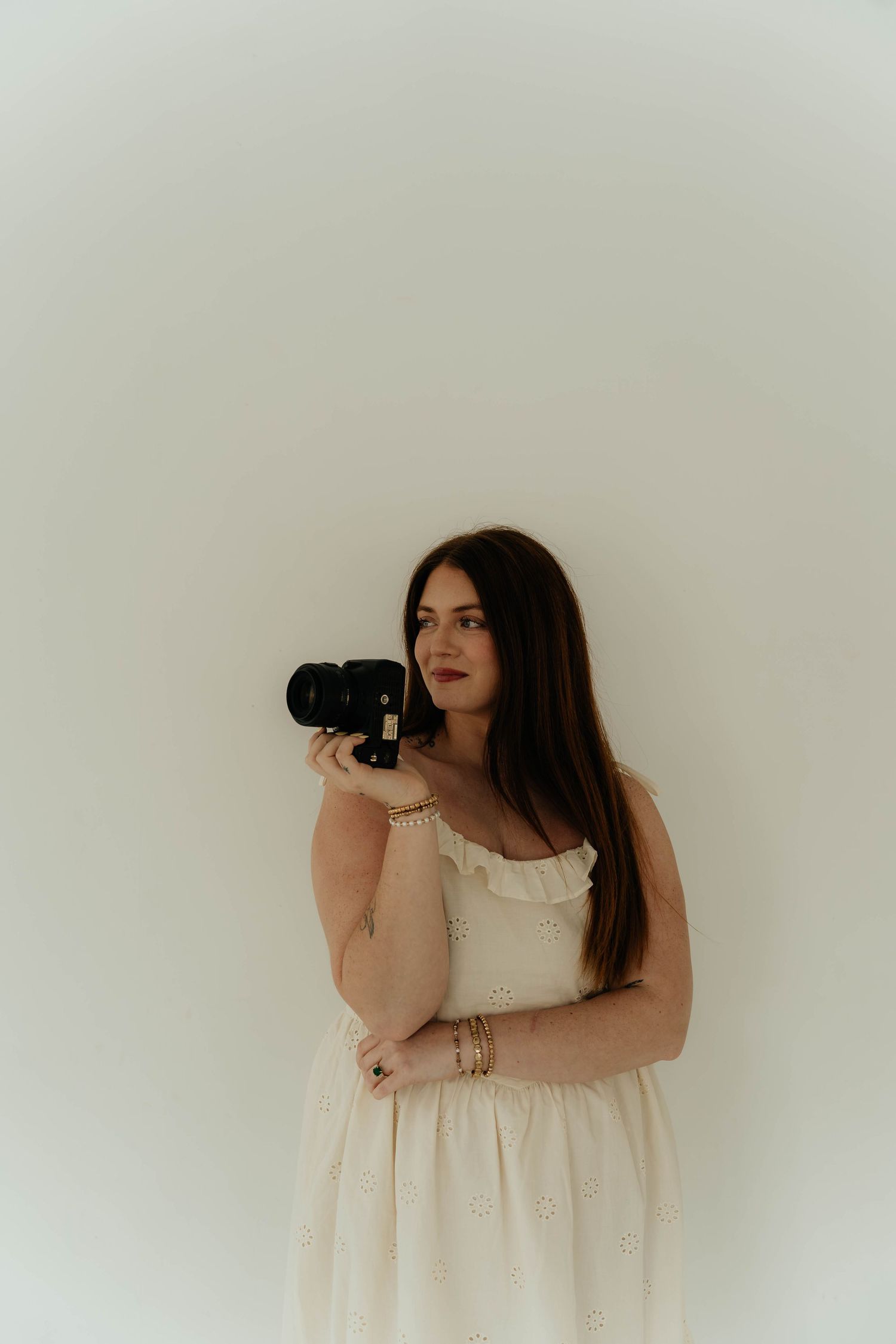 Woman in white dress holding a black camera, posing against a light background with long brown hair.