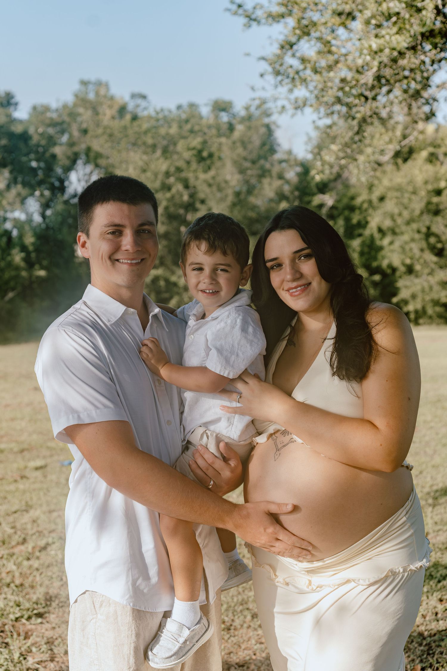 Smiling family maternity photo outdoors with father holding toddler son beside pregnant mother.