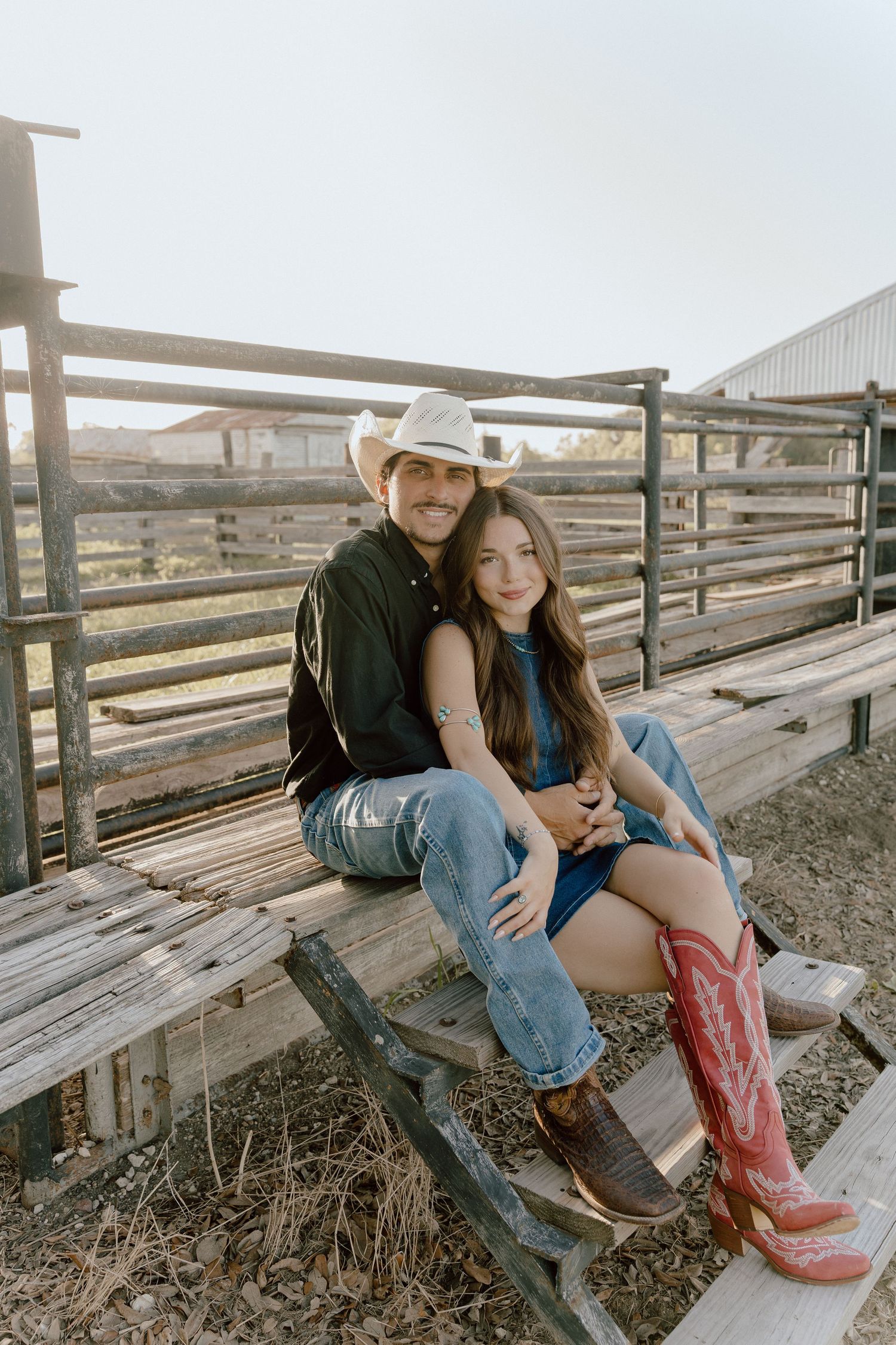 Couple sitting together on rustic wooden bleachers at a farm, woman wearing red cowboy boots and denim dress.