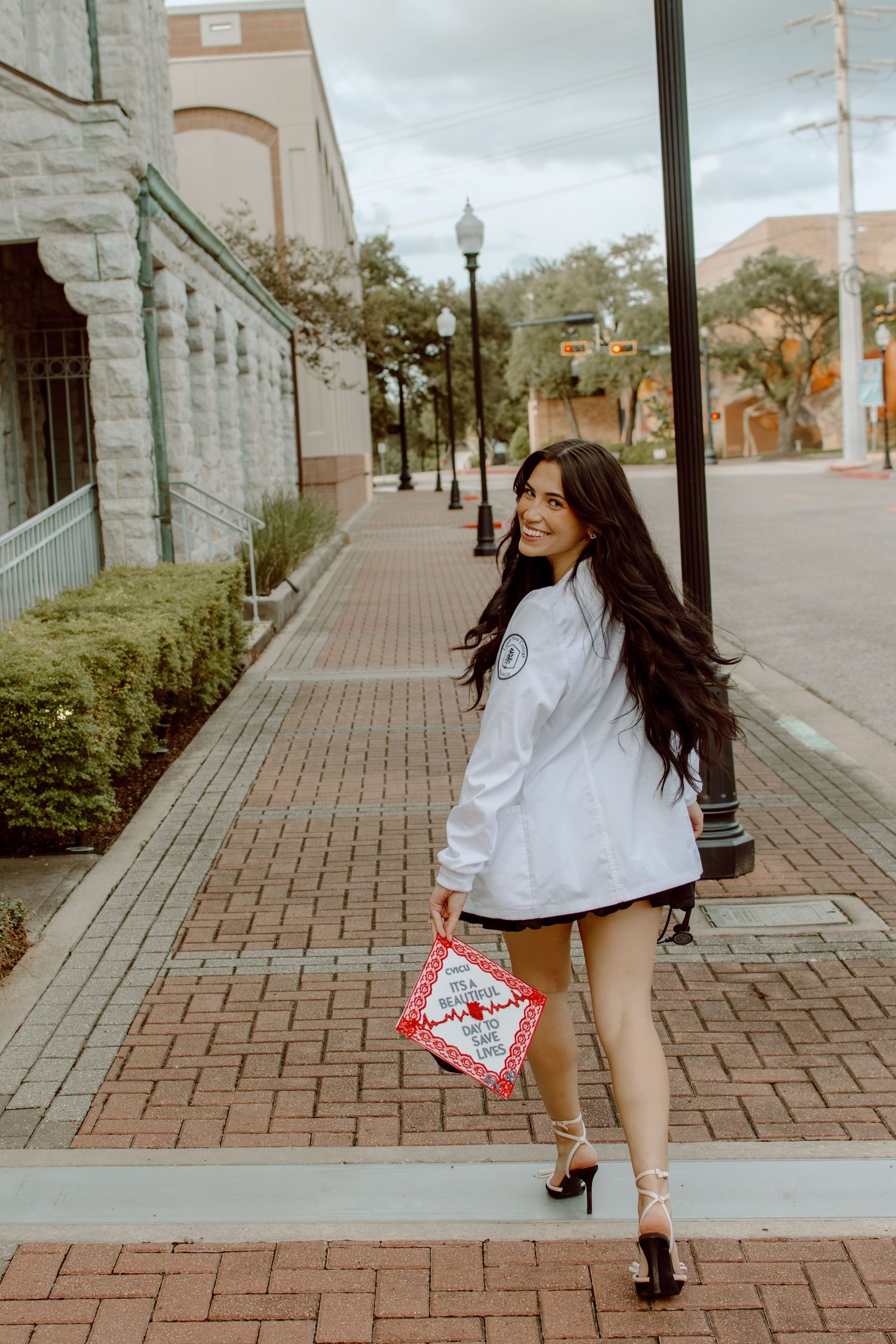 Smiling brunette woman walking on brick sidewalk holding graduation cap in white jacket.