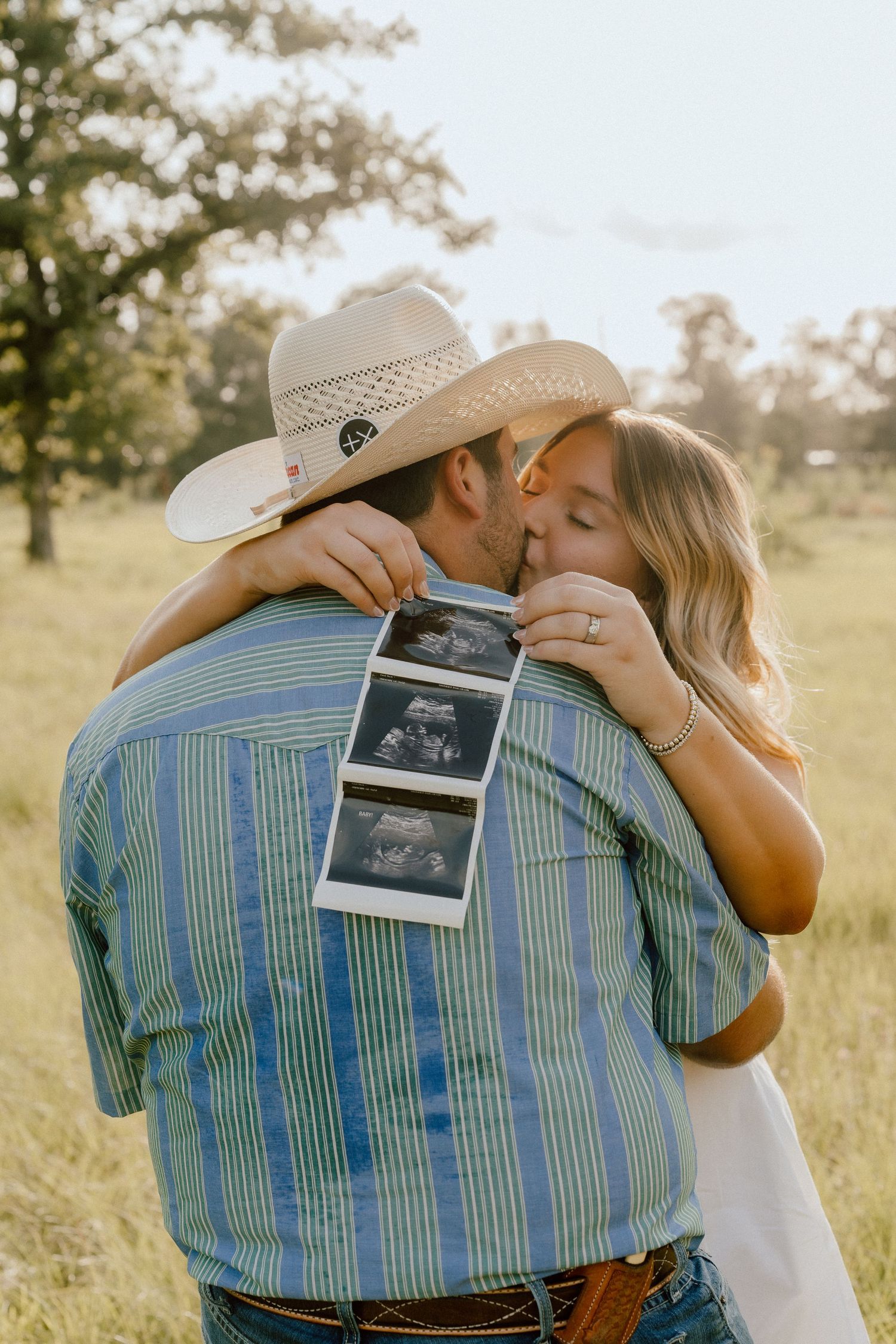 Couple kissing in a green field at sunset, woman holding ultrasound photos behind mans back as pregnancy announcement.