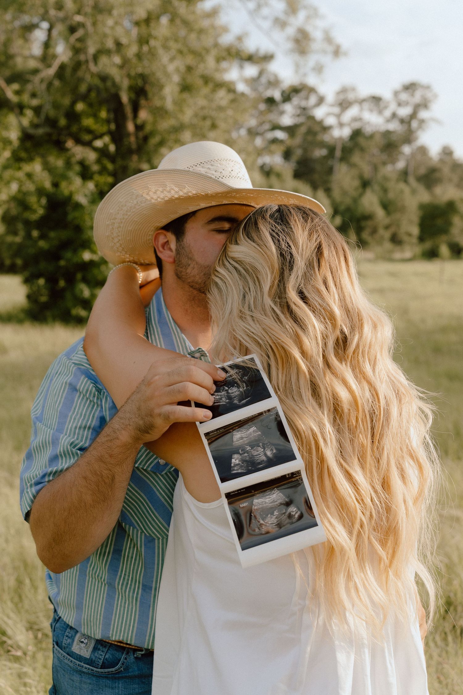 Couple kissing outdoors holding ultrasound photos for pregnancy announcement in a sunny field.