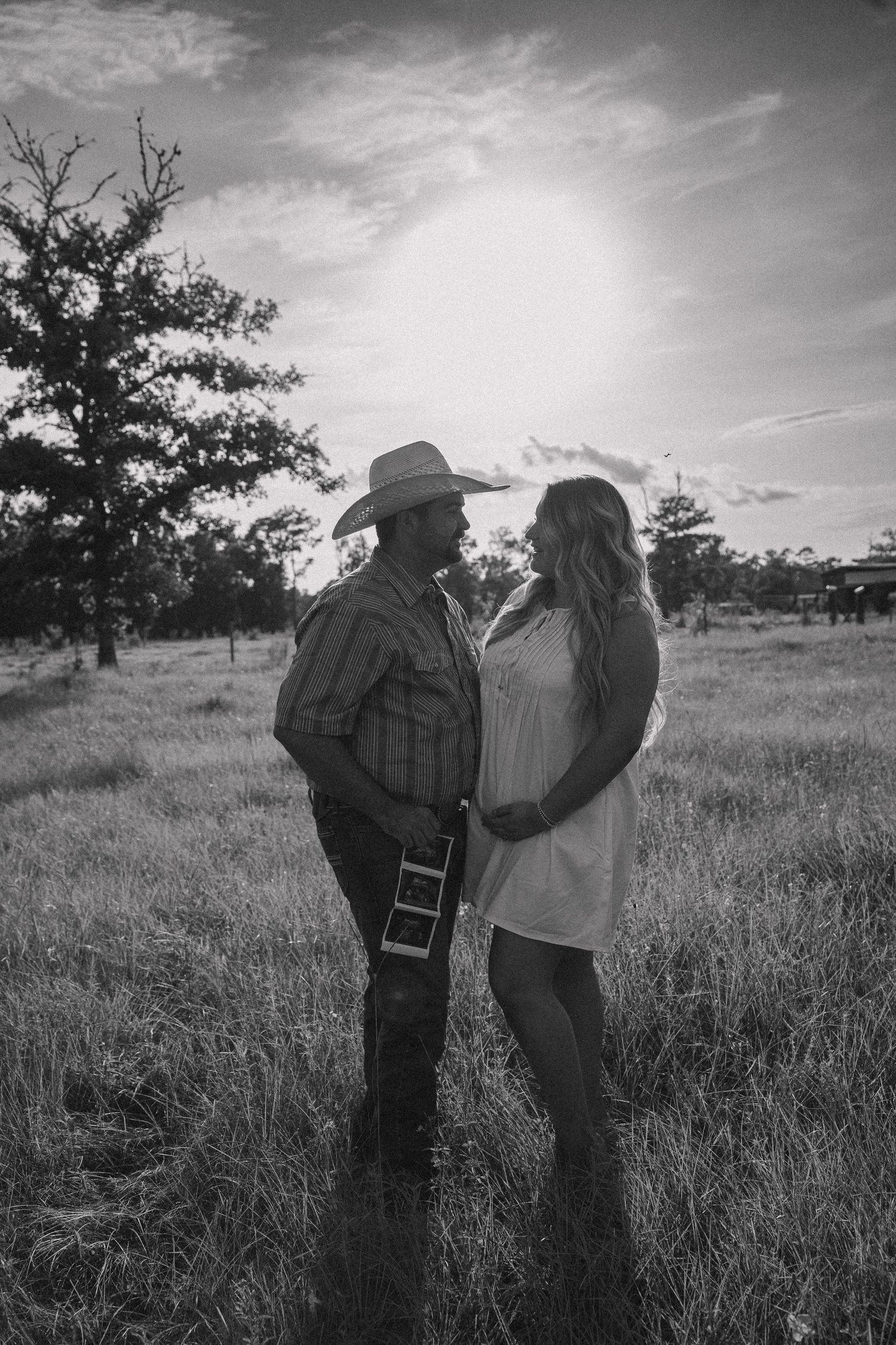 Black and white photo of expecting couple holding sonogram in sunlit open field.