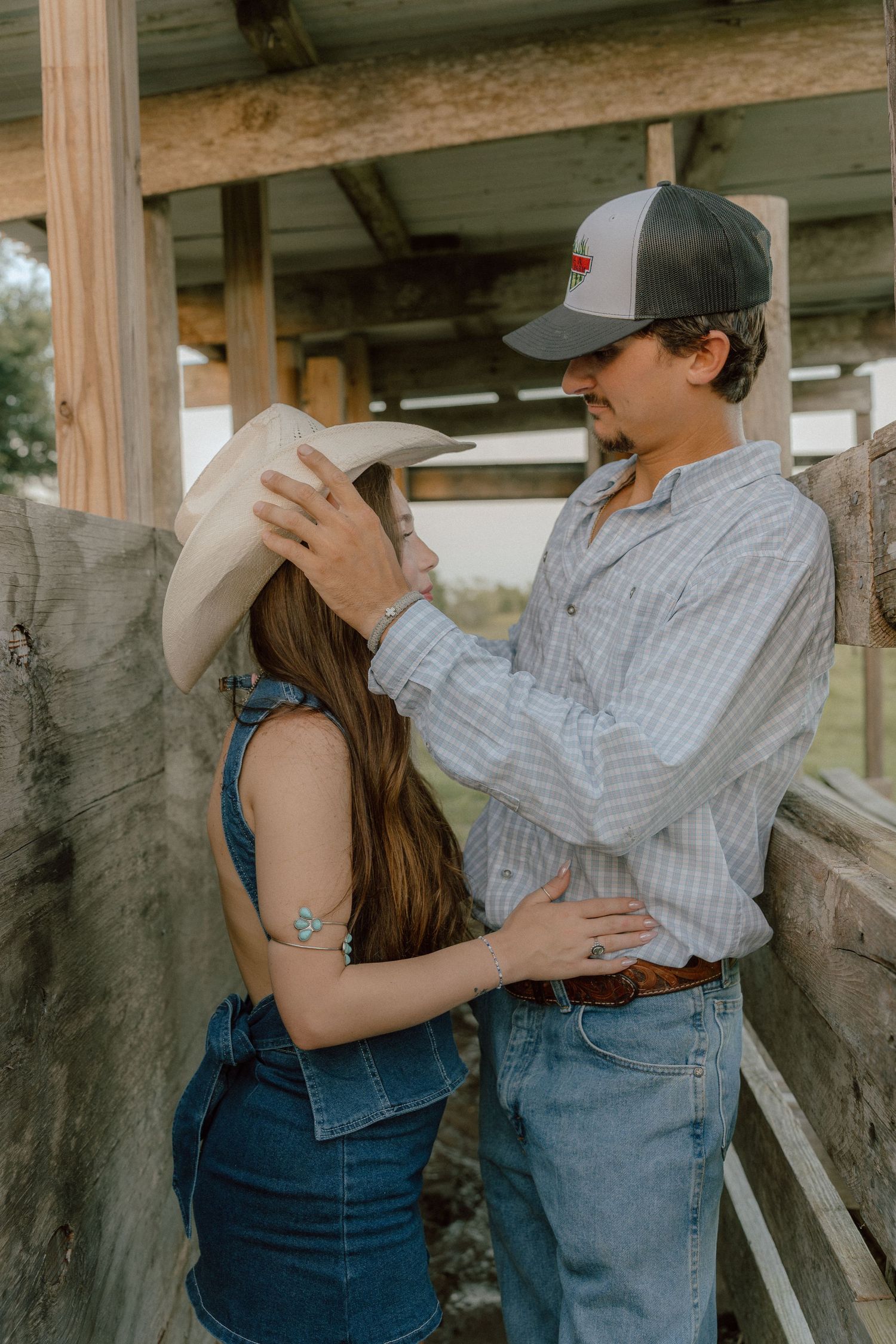 A man places a cowboy hat on a woman in denim overalls at a rustic wooden fence.