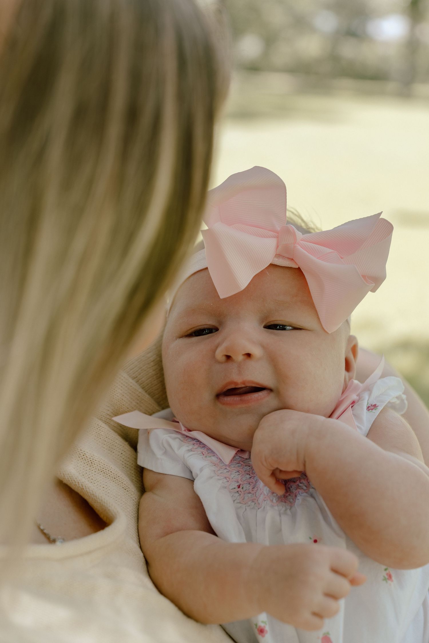 Newborn baby girl in pink bow headband and smocked dress being held by blonde mother outdoors.