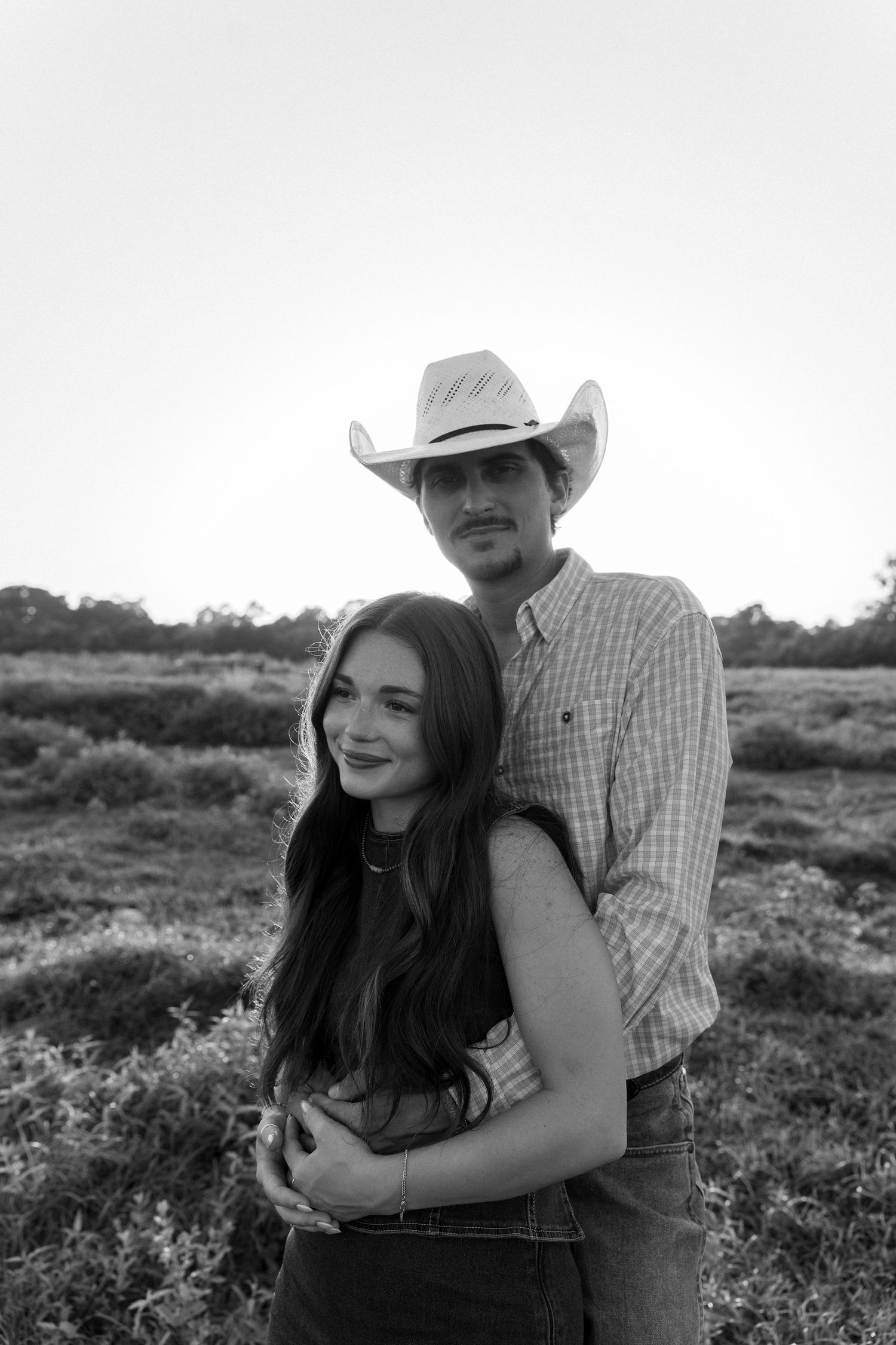 Black and white photo of couple embracing in open field at sunset, man wearing cowboy hat.