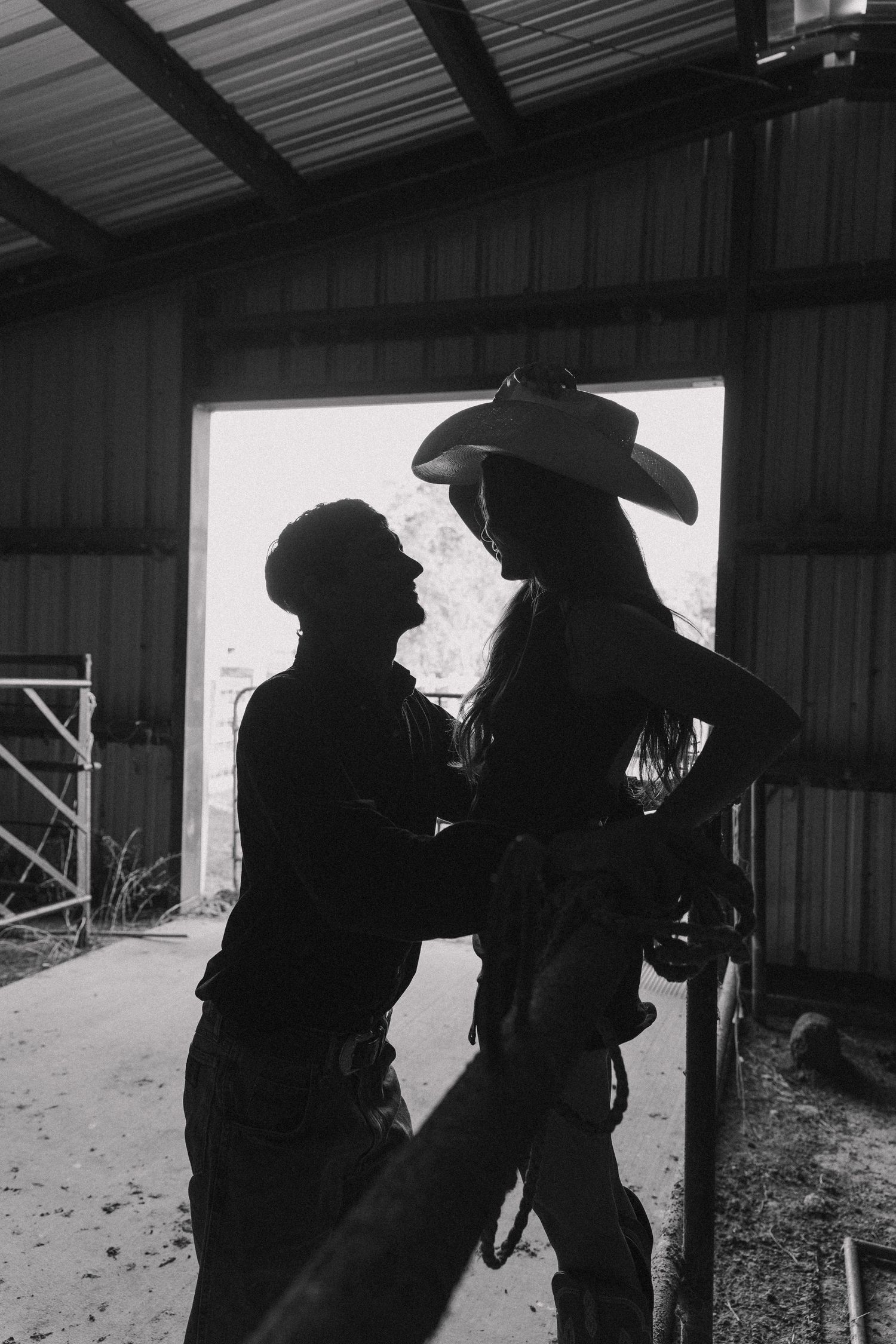 Black and white silhouette of couple sharing intimate moment in barn doorway, woman wearing cowboy hat.