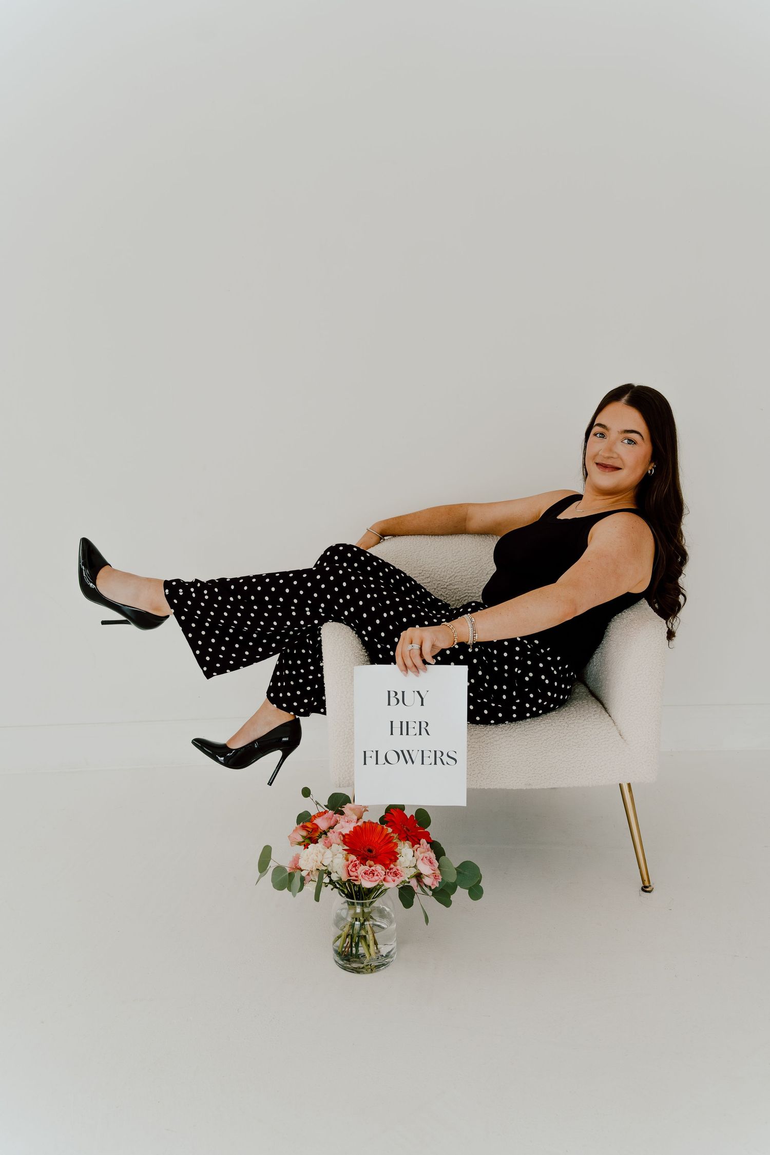 Woman in black polka dot pants sitting in white chair holding a Buy Her Flowers sign with red roses in foreground.