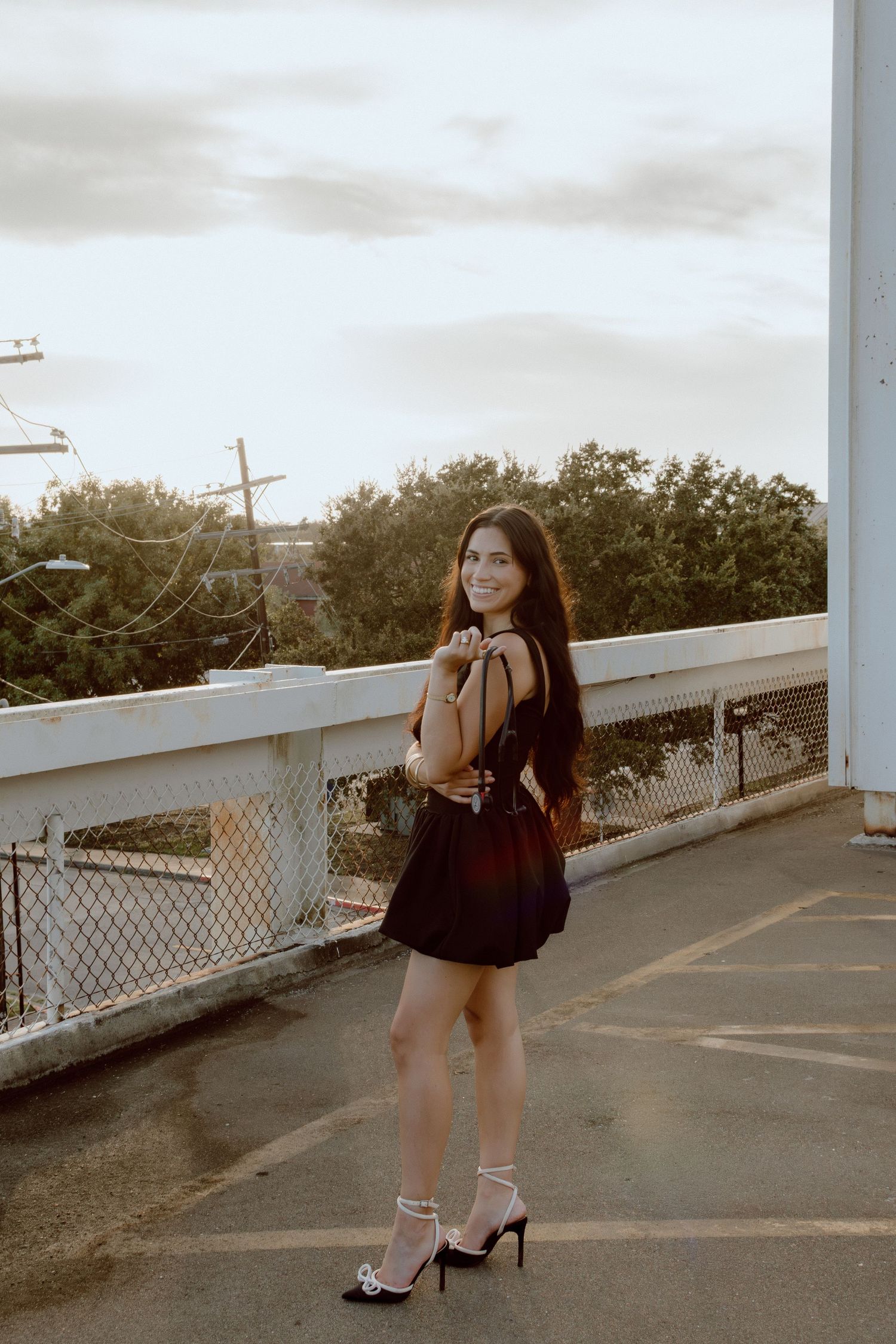 Young woman in black mini dress and heeled sandals smiling on a rooftop parking deck.