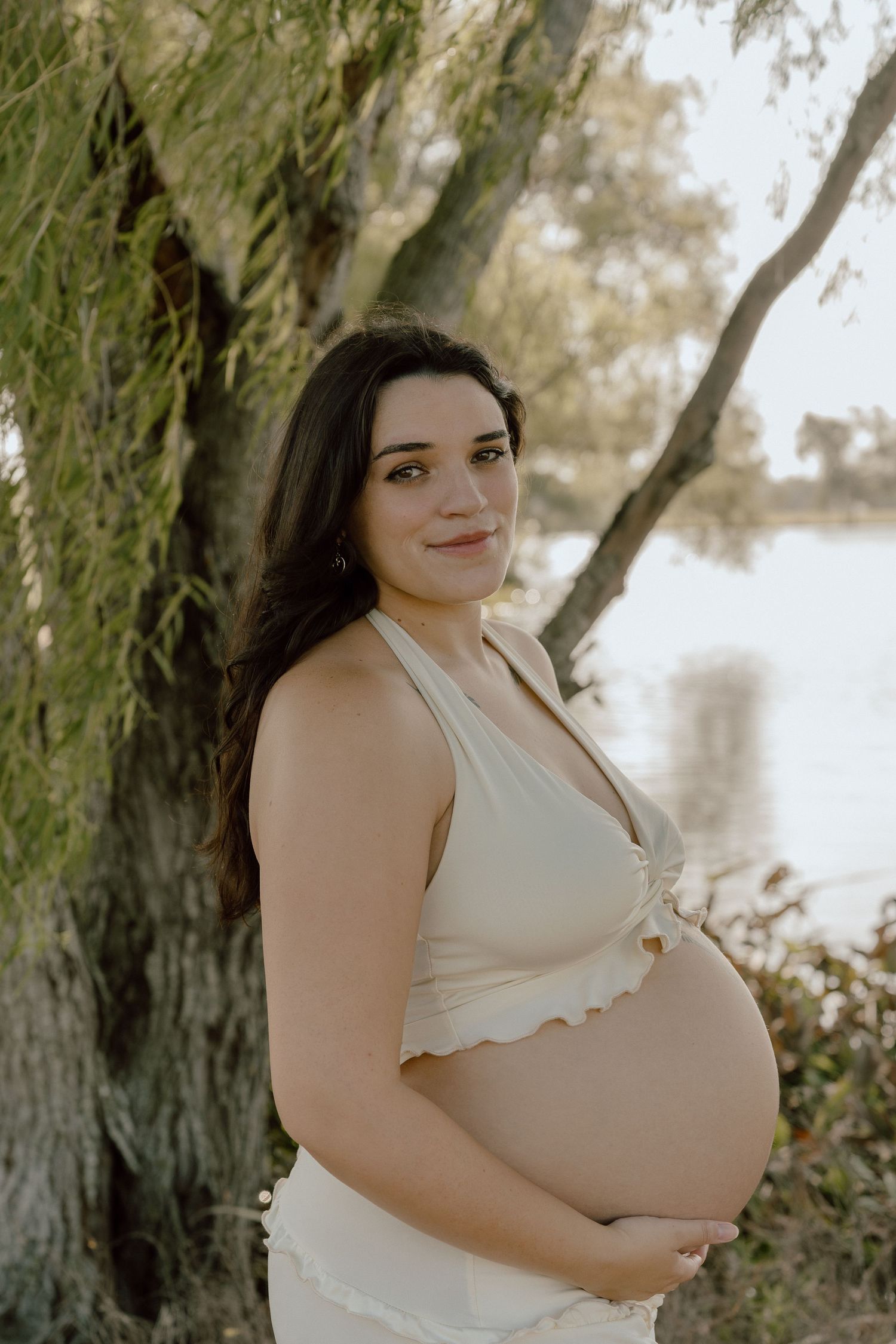 Pregnant woman in a cream crop top cradling her baby bump near a lakeside tree.