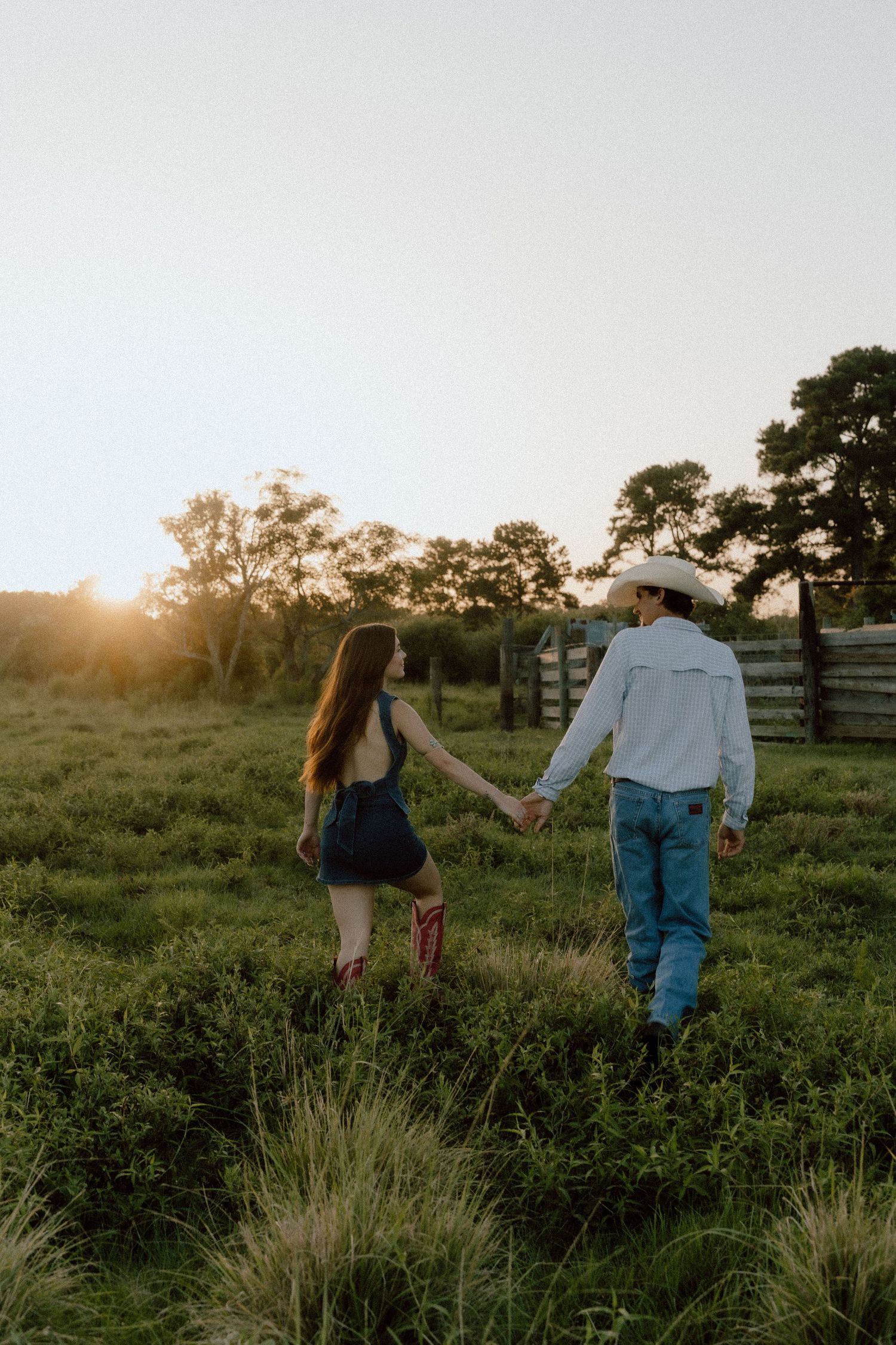 Couple holds hands walking through green field at golden sunset near rustic wooden fence.