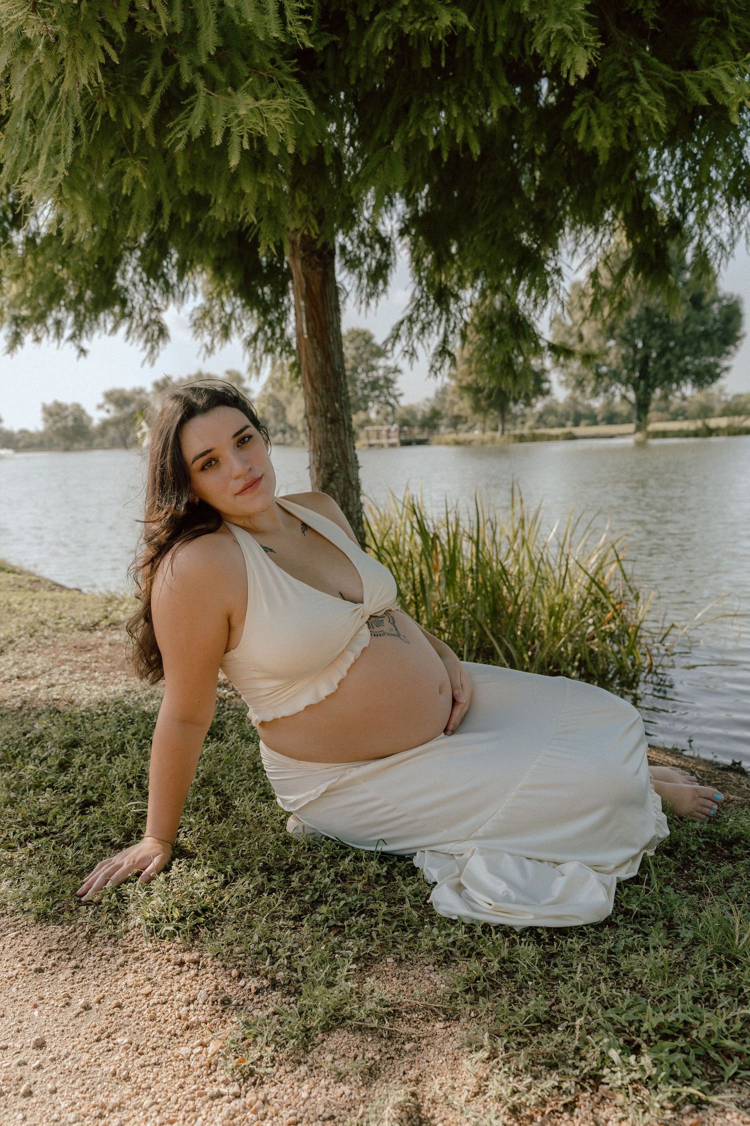 Pregnant woman in cream crop top and skirt sits gracefully under tree beside tranquil lake.