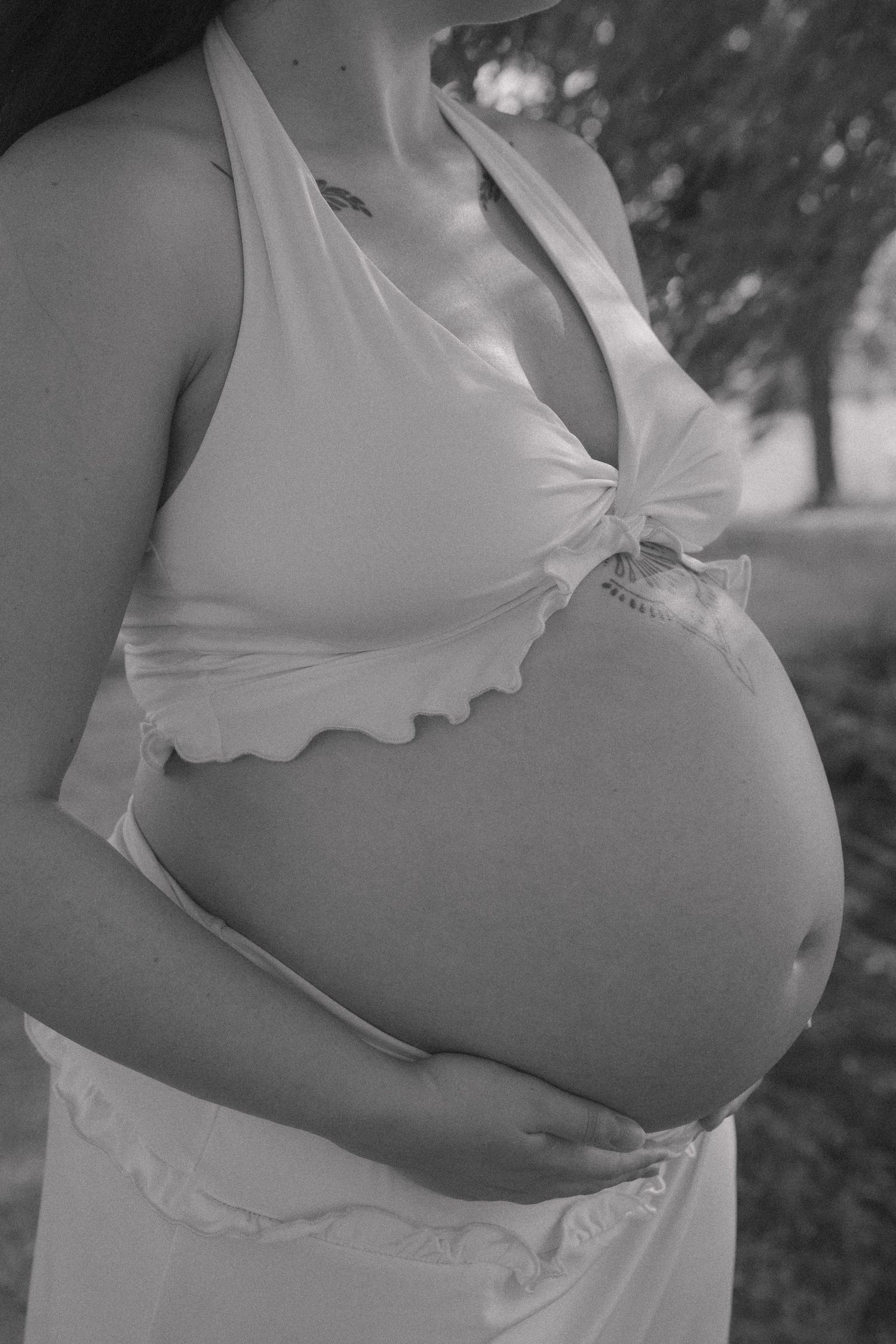 Black and white close-up of pregnant woman in white halter top cradling baby bump outdoors.
