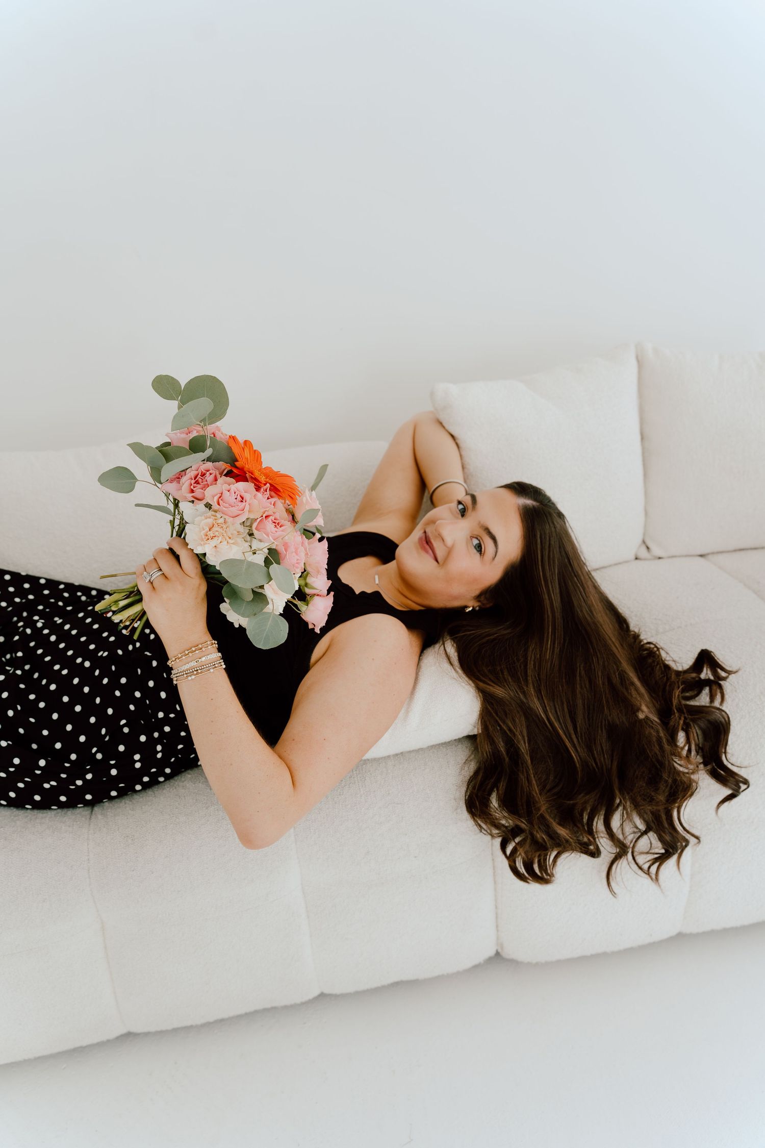 Young woman with long dark hair lying on white sofa holding a colorful bouquet of flowers.