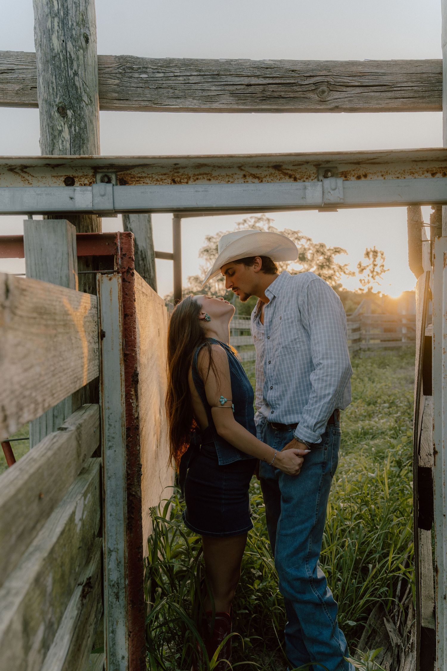 Couple sharing a romantic moment in rustic farm gate at golden hour, man in cowboy hat.