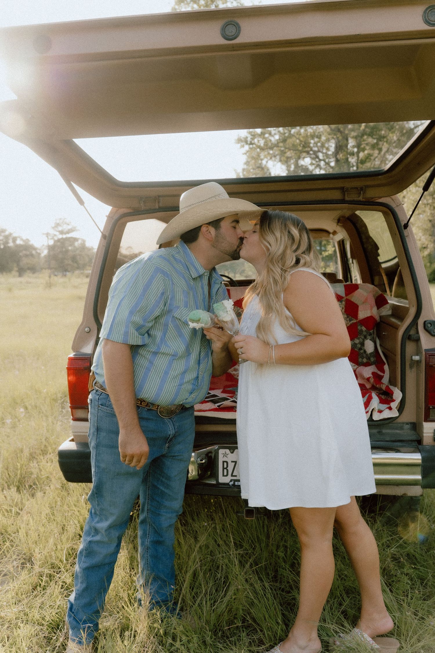 Couple kissing in golden sunlight by vintage SUV tailgate in open grassy field.