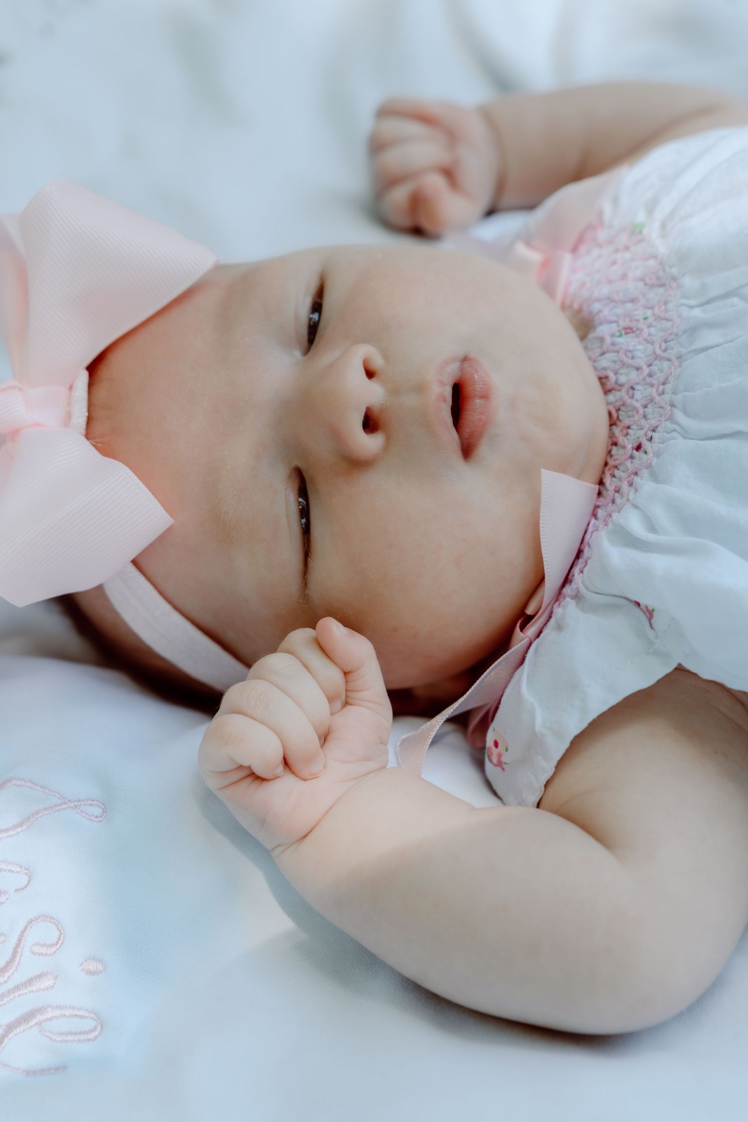 Newborn baby girl in pink bow headband lying on white blanket with tiny fists raised near her face.
