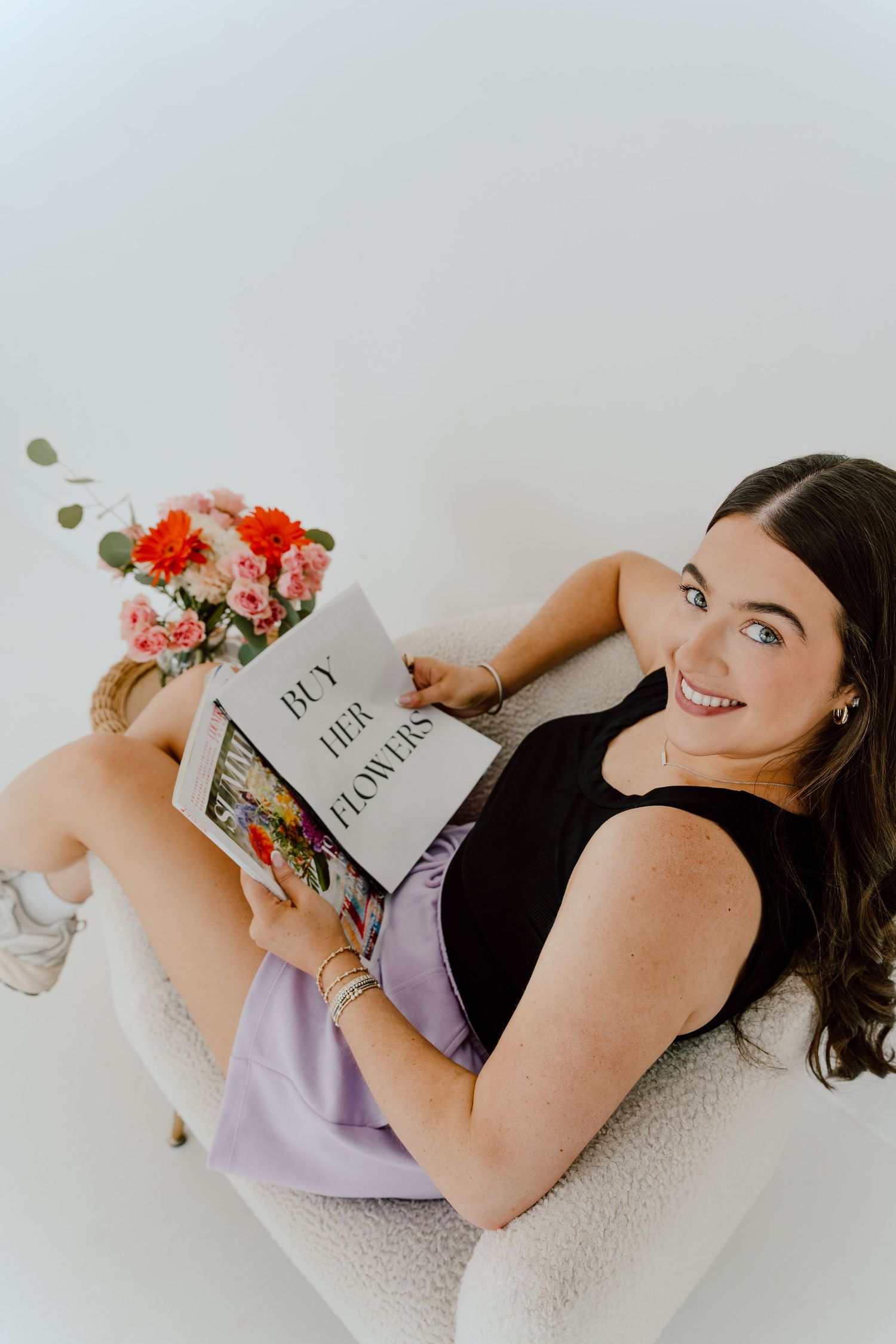 Smiling woman sitting in white chair holding a book titled Buy Her Flowers with floral arrangement nearby.