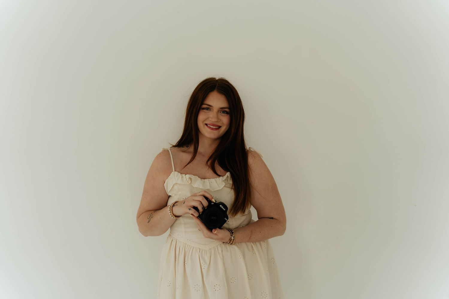Smiling young woman in a cream dress holding a camera, posing against a white background.
