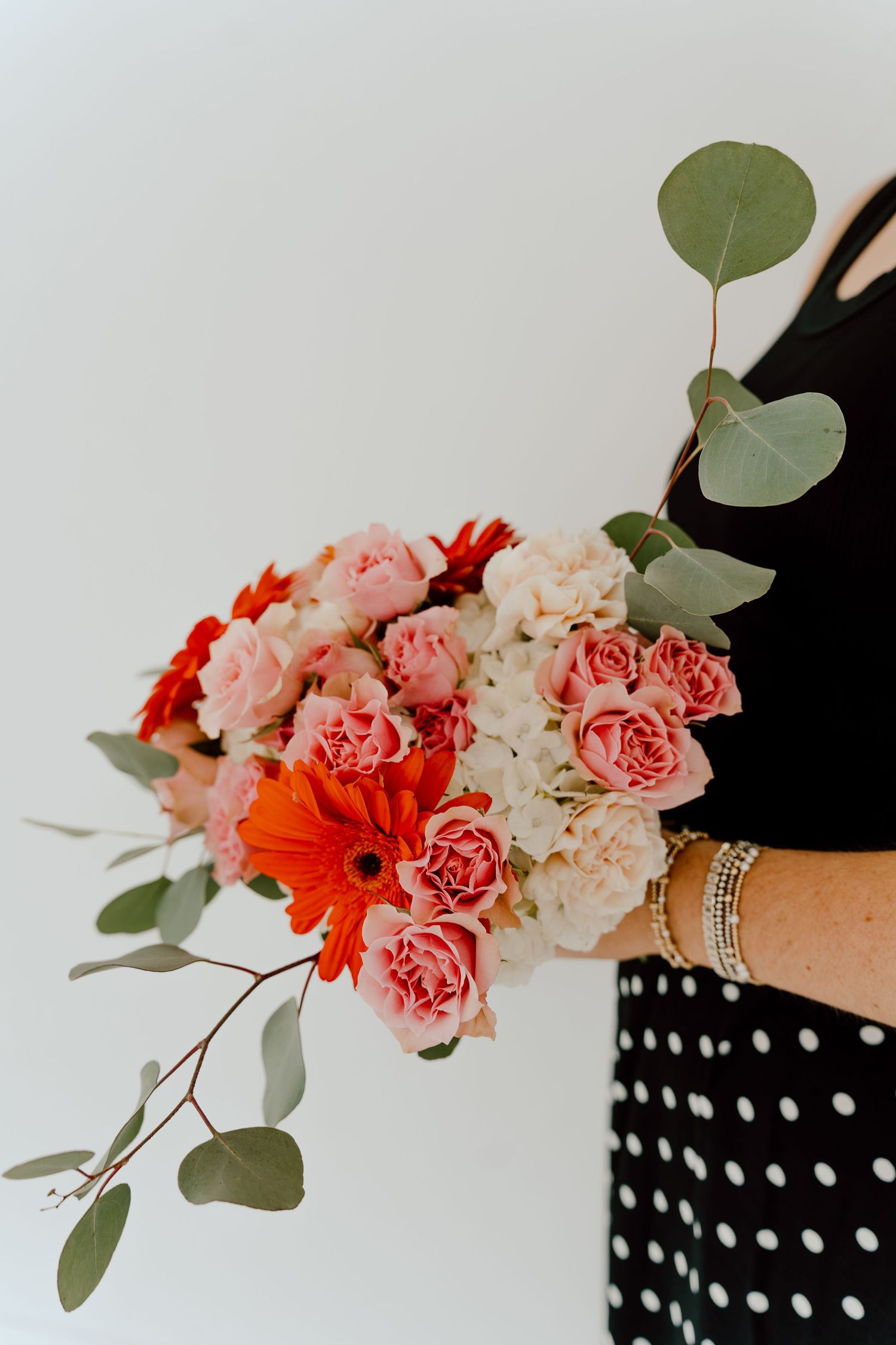 A woman in a black polka dot dress holds a lush bouquet of pink roses, red gerbera daisies, and eucalyptus stems.