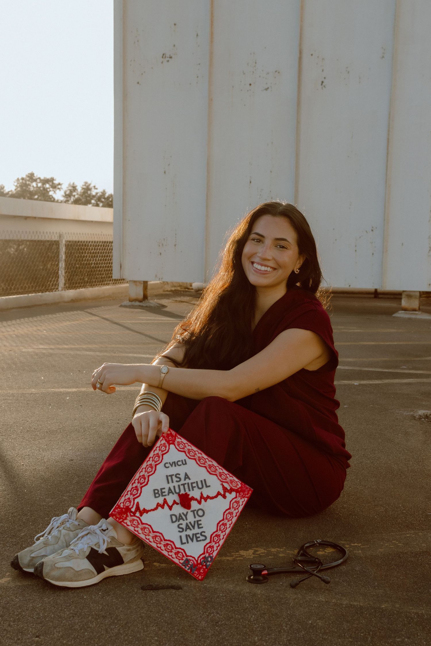 Nursing graduate in burgundy scrubs sitting with decorated cap reading it's a beautiful day to save lives.