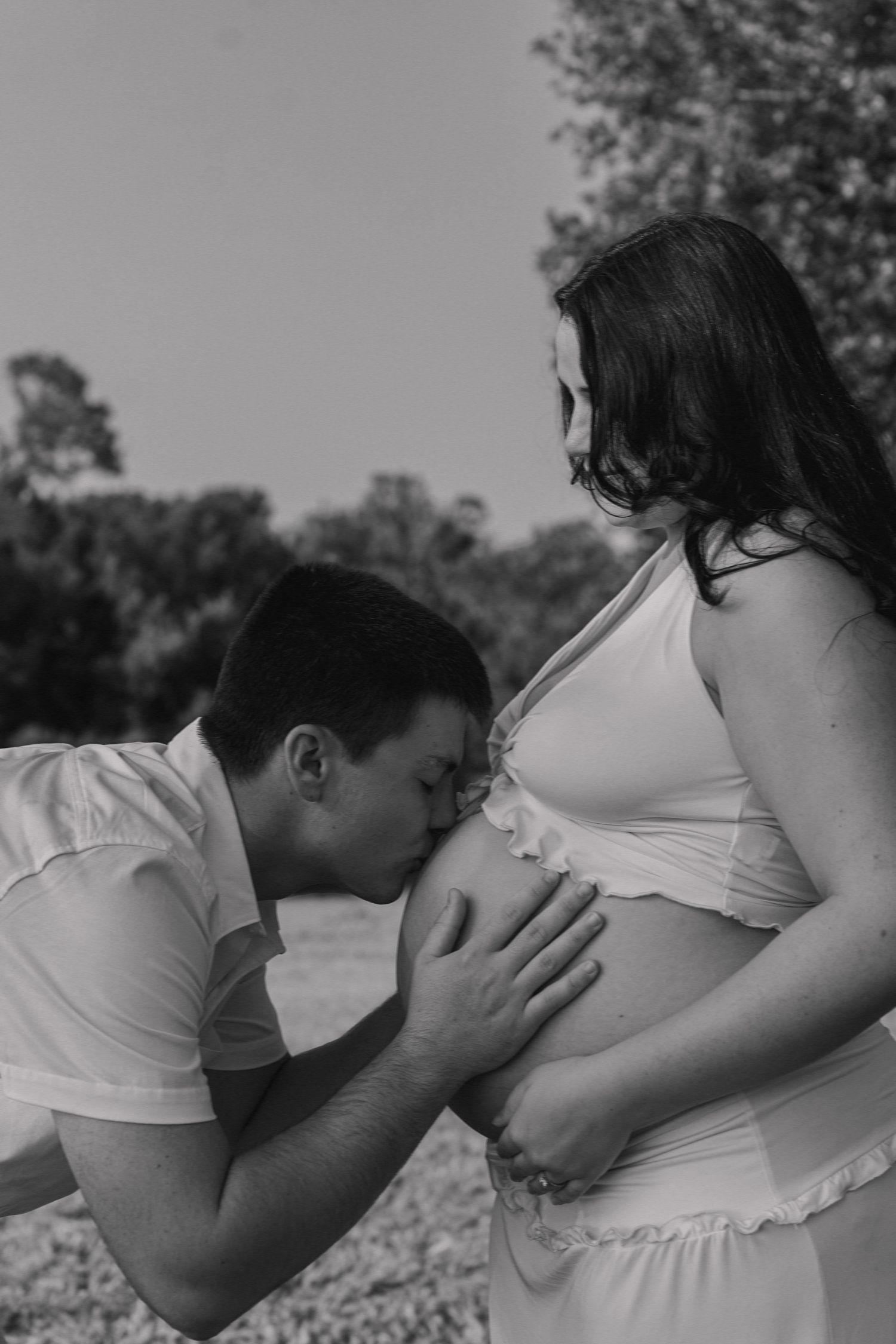 Black and white photo of a man kissing a pregnant woman's baby bump outdoors.