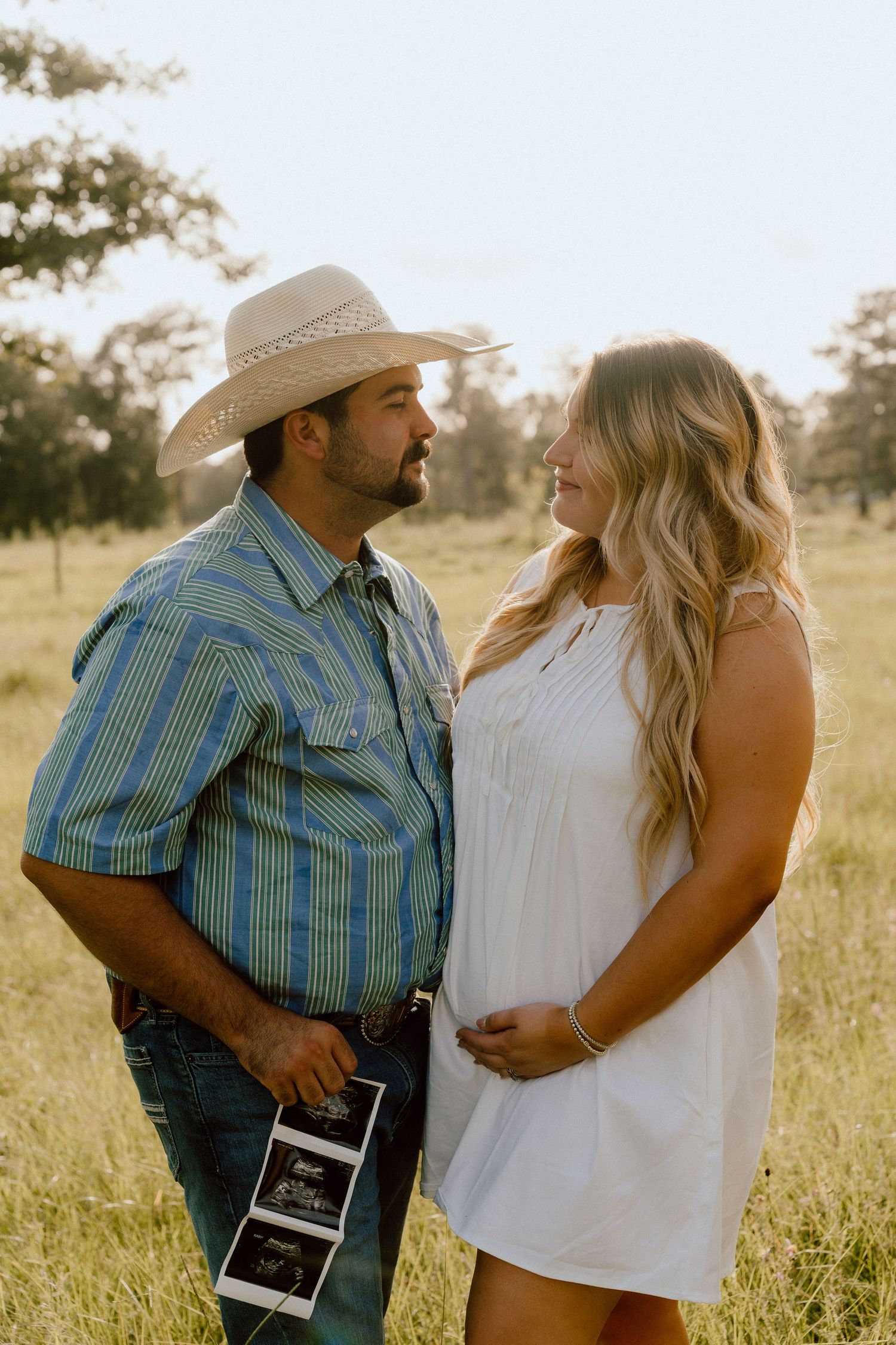 Cowboy and pregnant woman in white dress sharing a tender moment in a sunny field.